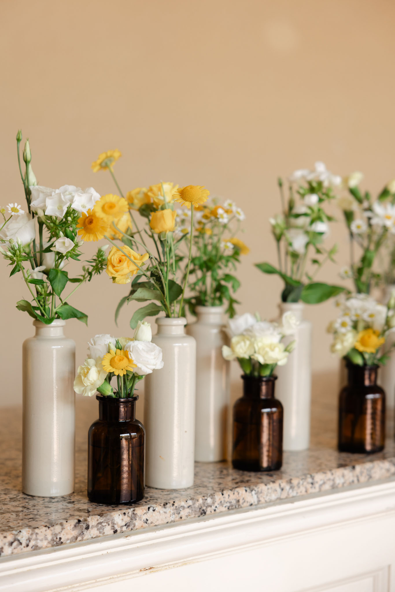 Bud vases with yellow roses, craspedia, and white ranunculus arranged on a granite mantelpiece