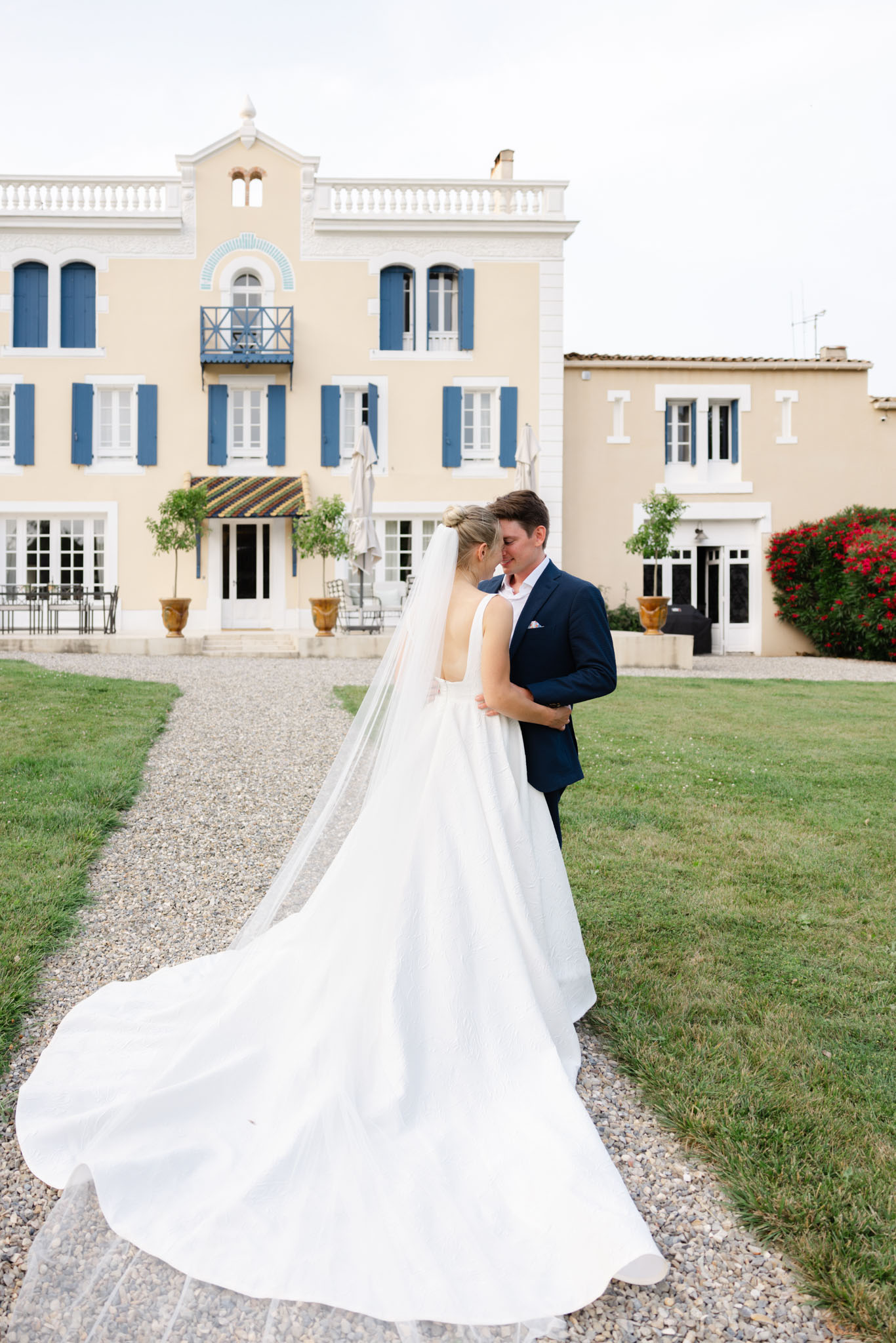 Bride and groom touching foreheads on gravel path in front of yellow Provencal manor with blue shutters