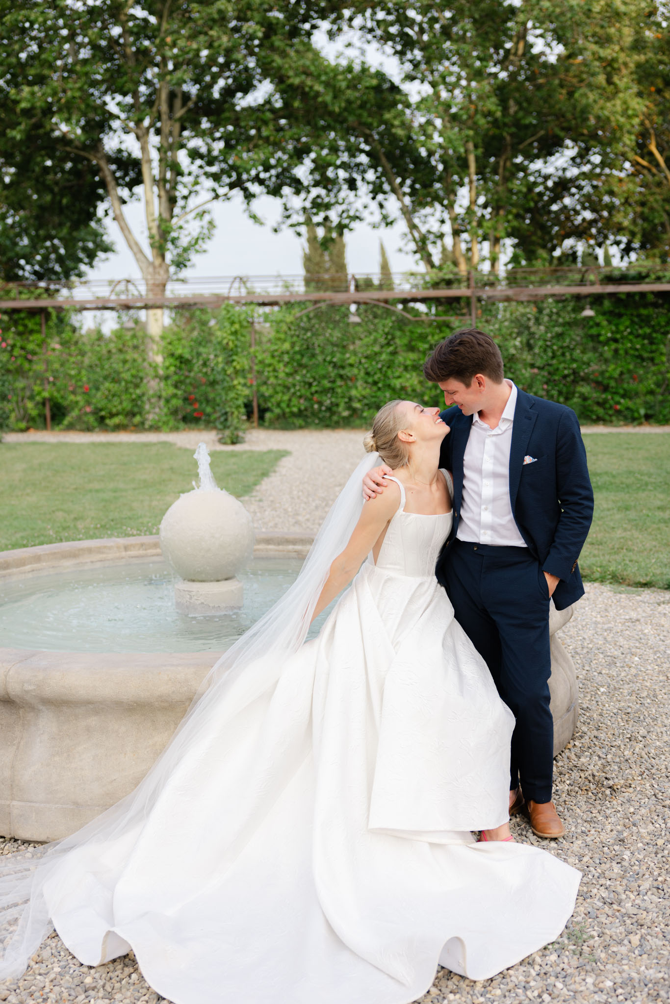 Bride in white ballgown with cathedral veil and groom in navy suit laughing beside stone garden fountain