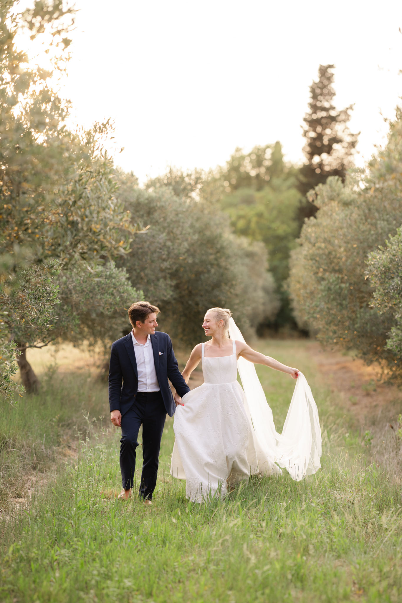 Bride in white A-line gown with cathedral veil and groom in navy suit walking through olive grove