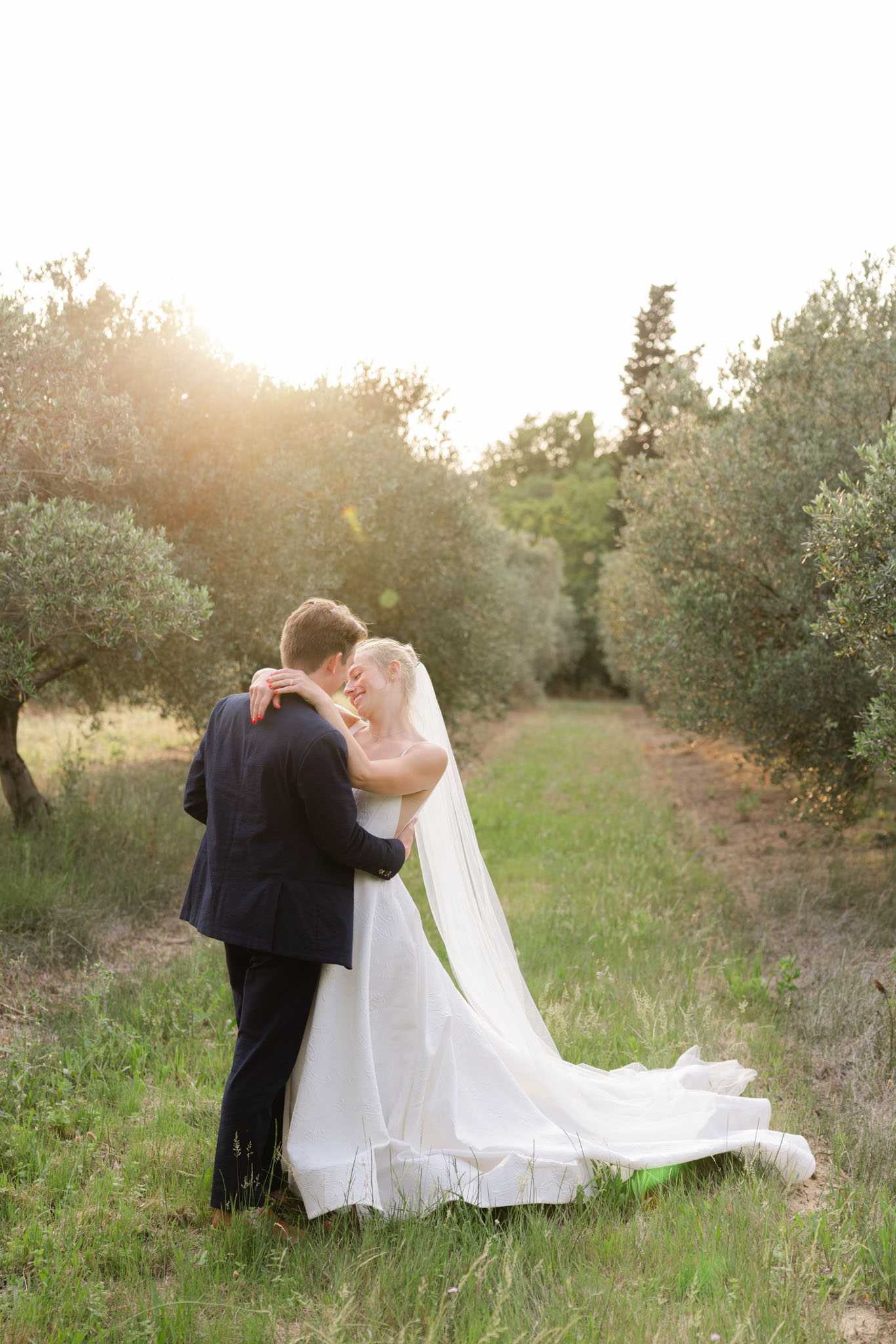 Bride in white strapless ball gown and cathedral veil embracing groom in navy suit in an olive grove at golden hour