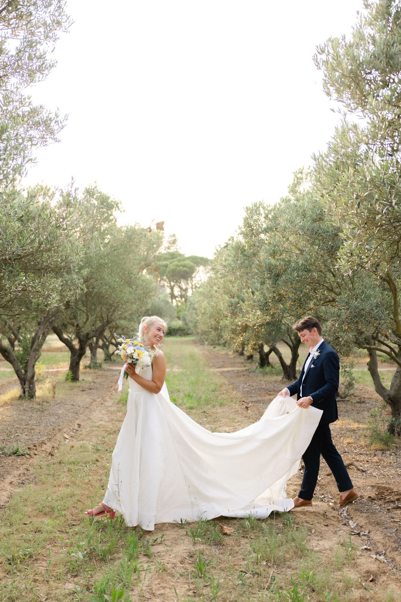 Groom carries bride's train as they walk through an olive grove, bride holding wildflower bouquet