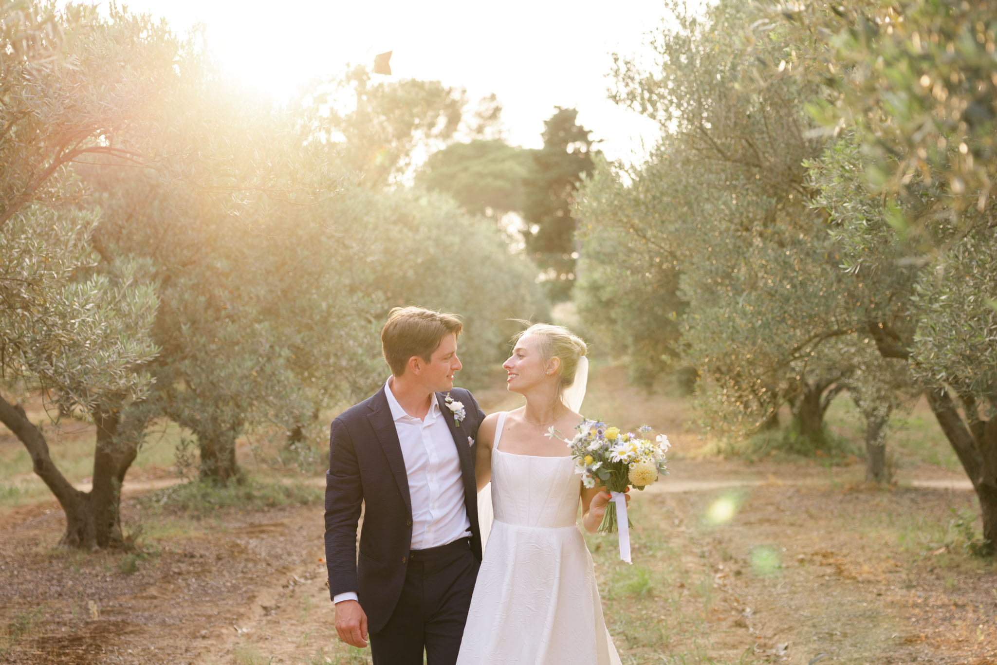 Bride and groom walking through olive grove at golden hour, bride carrying wildflower bouquet with daisies
