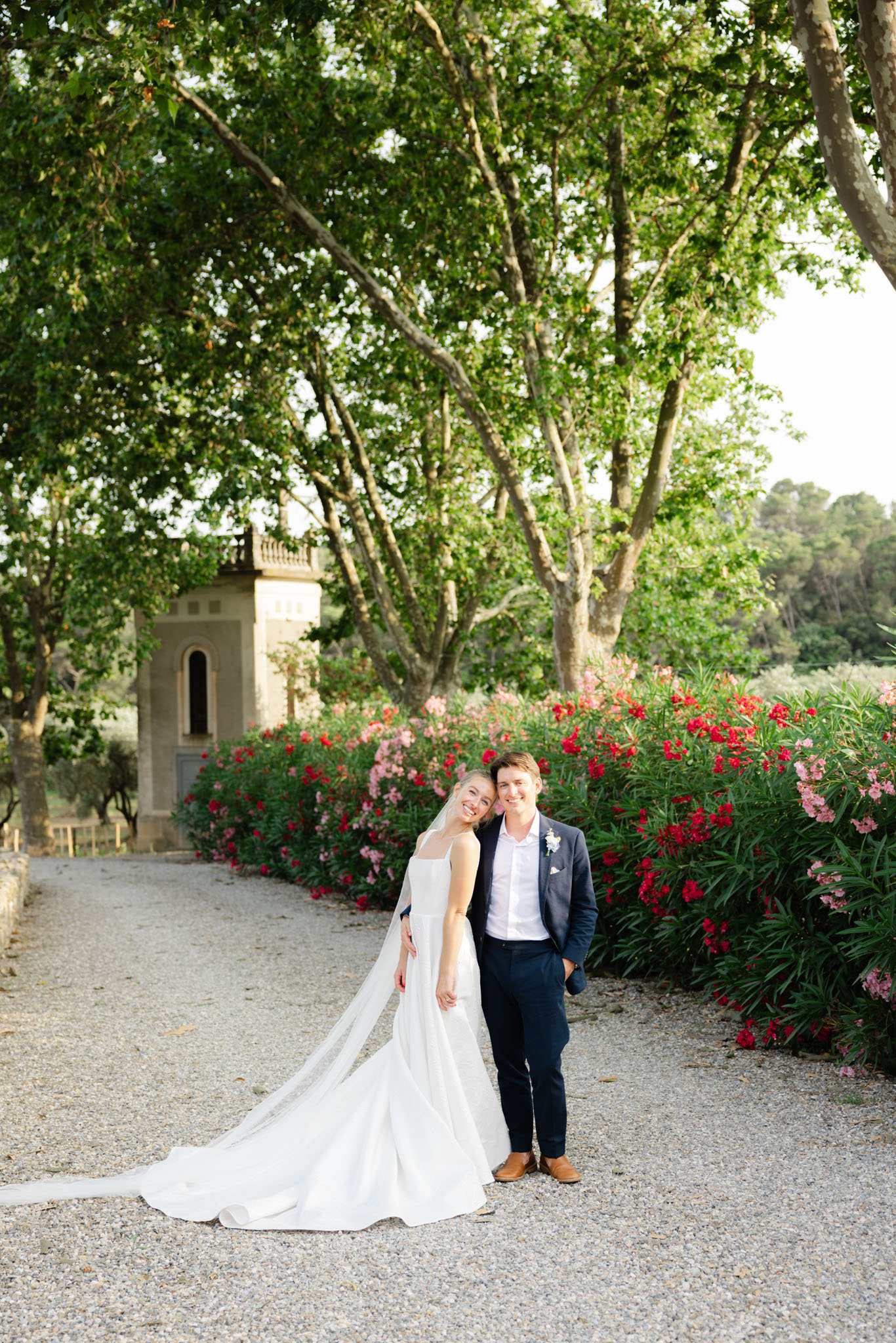 Couple on gravel path bride in sleeveless gown with cathedral train and veil groom in navy suit red oleander behind