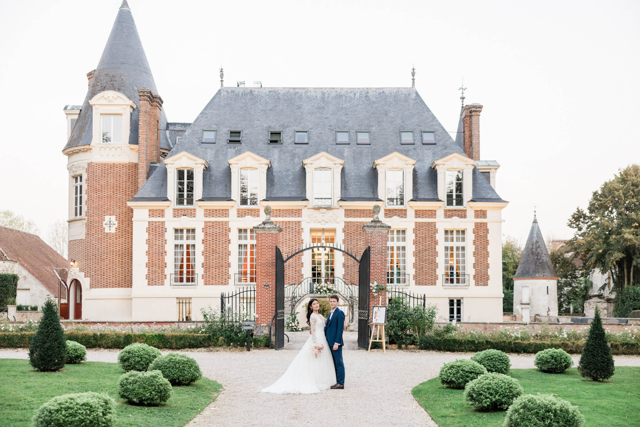 A couple poses for a portrait on the gravel pathway leading to the entrance of a French château, with the full façade of the building visible behind them. The bride wears a long-sleeved lace ball gown in ivory with a cathedral-length train and carries a bouquet with soft pink and white blooms, while the groom wears a navy blue suit. The château is a multi-story red brick and cream stone building with a dark slate mansard roof, conical corner towers, wrought-iron entrance gates decorated with floral arrangements, and warmly lit interior windows visible through the gate arch. A wooden easel with a printed wedding sign stands to the right of the couple near the gate. The foreground garden features neatly trimmed spherical boxwood topiaries and conical yew shrubs lining the pathway. This is a wide shot taken at dusk in a classic French château style. Potential venue feature image.
