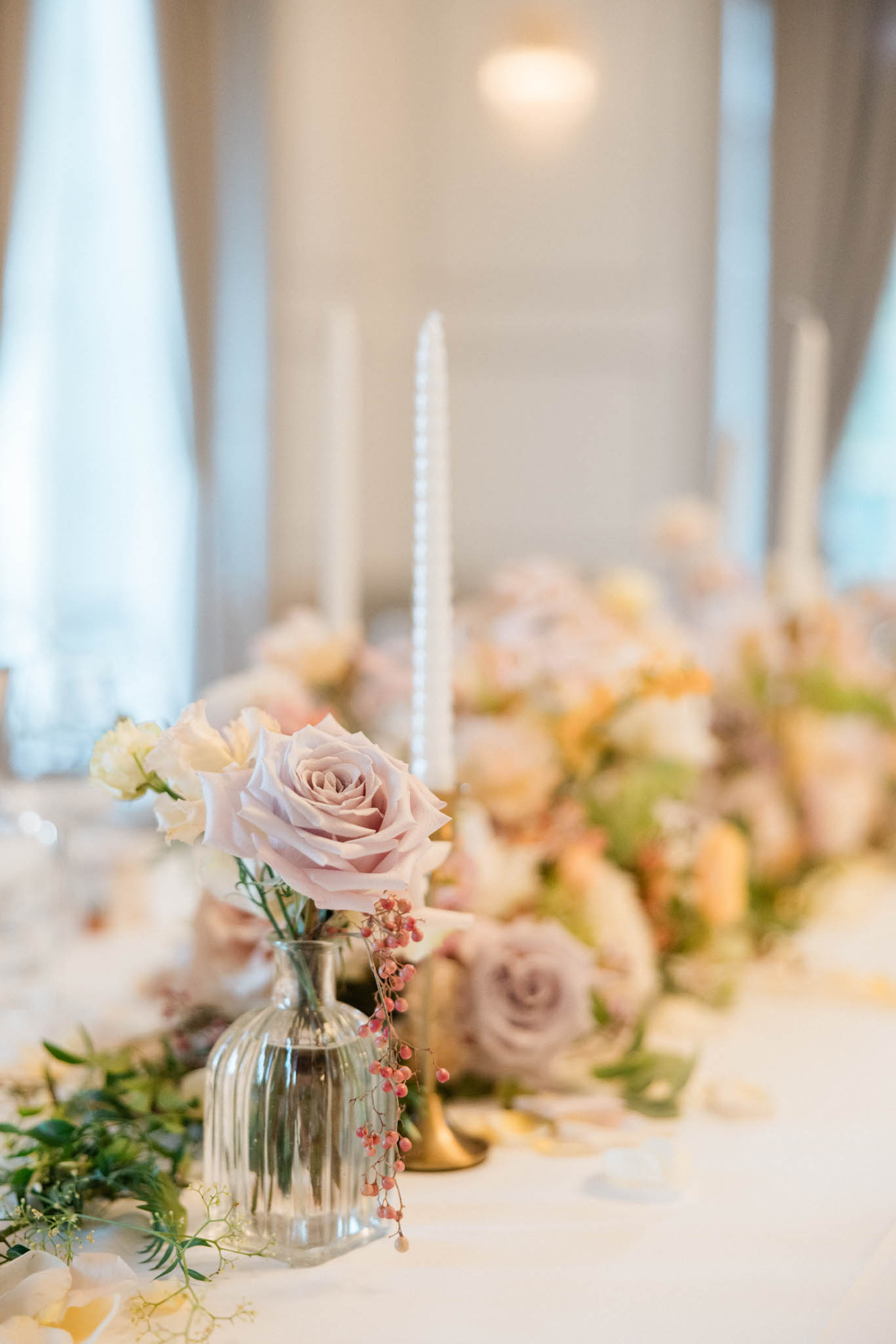 Ribbed bud vase with dusty mauve rose and pepper berries beside gold candlestick on white linen