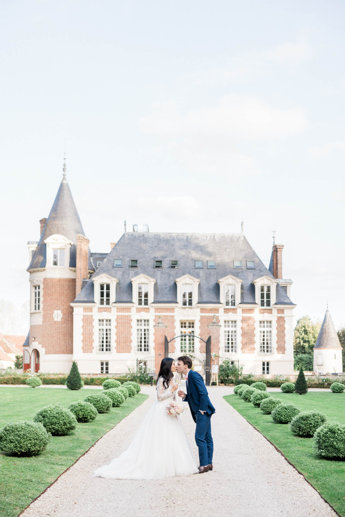 Couple kissing on topiary-lined gravel path before red brick and white stone chateau with mansard roofs