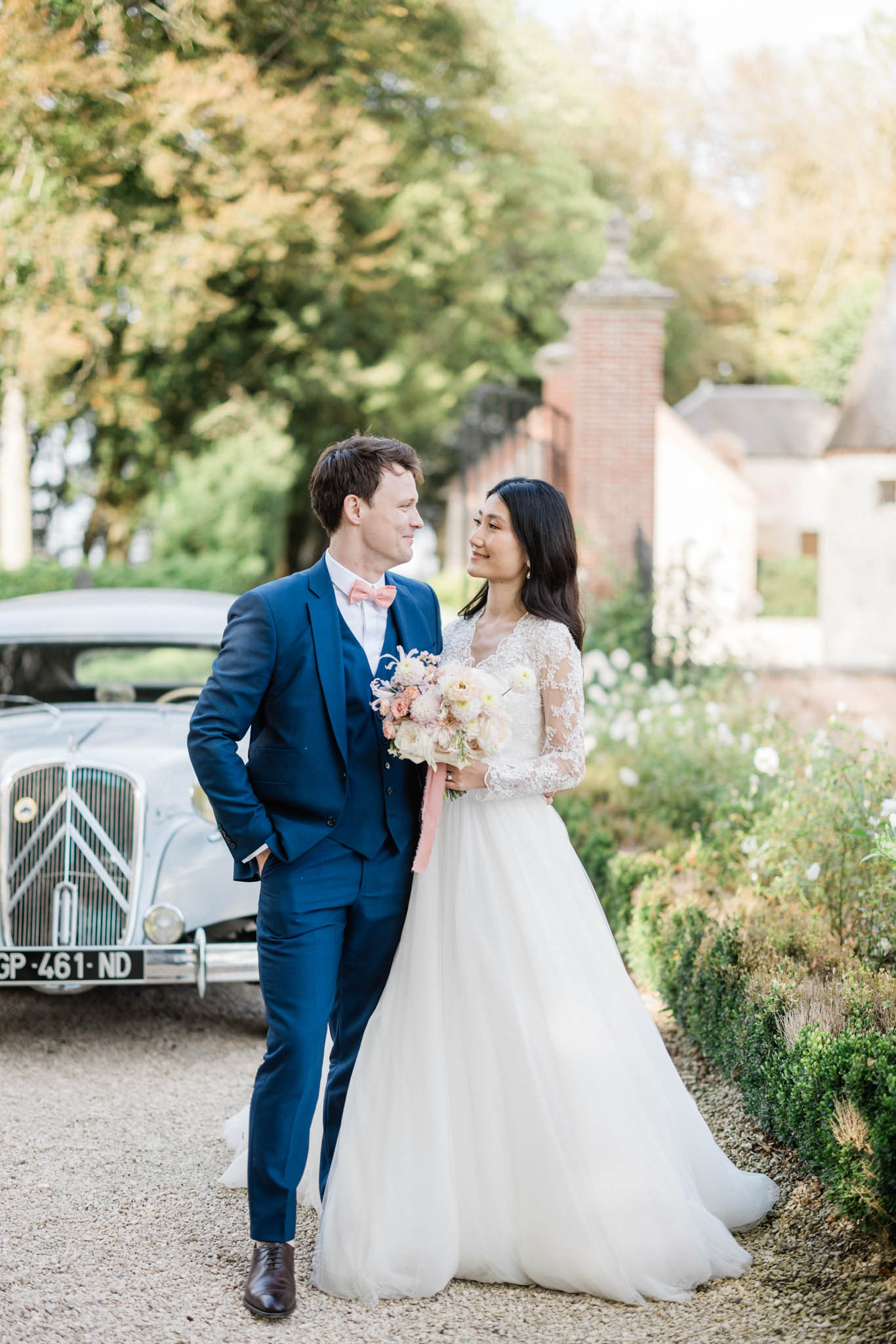 A couple portrait shot outdoors on a gravel driveway, with the bride and groom looking at each other and smiling. The groom wears a navy blue three-piece suit with a salmon-pink bow tie and brown leather Oxford shoes. The bride wears a long-sleeve lace-bodice ballgown with a full tulle skirt, and carries a loose bouquet of blush and cream blooms — including what appear to be garden roses and dahlias — tied with a draped dusty pink ribbon. A vintage Citroën traction avant in pale grey is parked in the background, along with brick gate pillars and a stone outbuilding partially visible to the right. The styling palette combines navy, blush, and ivory in a classic French-countryside aesthetic. Medium full-length portrait composition.
