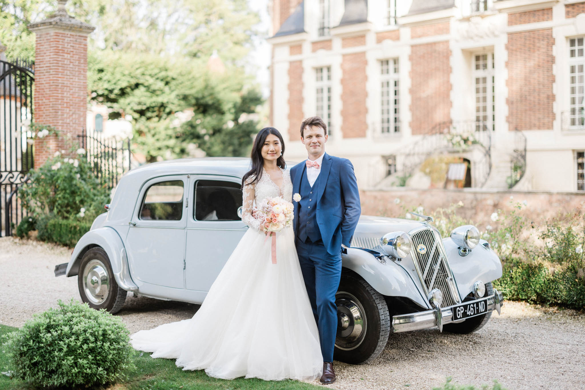 Couple before pale blue vintage Citroen at chateau bride in lace-sleeve ballgown with blush and coral dahlia bouquet