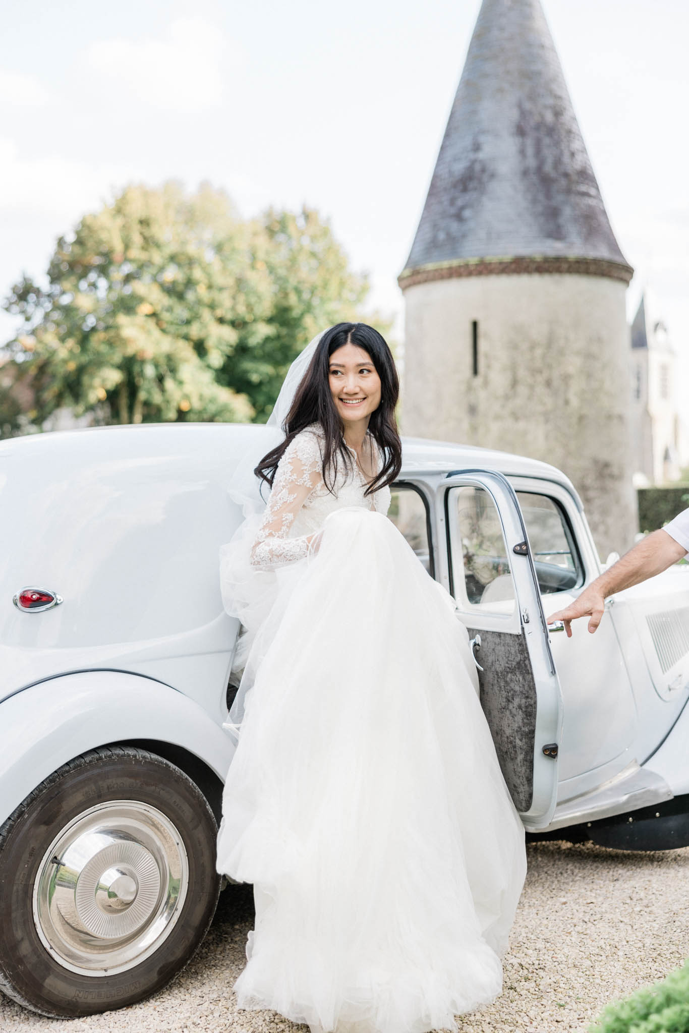 A bride steps out of a white vintage car, likely a Citroën 2CV or similar classic French vehicle, on a gravel driveway outside a château with a distinctive stone turret visible in the background. She is wearing a white ball gown with a long-sleeve lace illusion bodice and a full voluminous tulle skirt, paired with a cathedral-length veil, and she turns to smile toward the camera. A hand, presumably the groom's, holds the car door open for her. The portrait-style shot is taken outdoors in natural daylight, with a classic French château setting and a romantic yet unfussy styling approach.