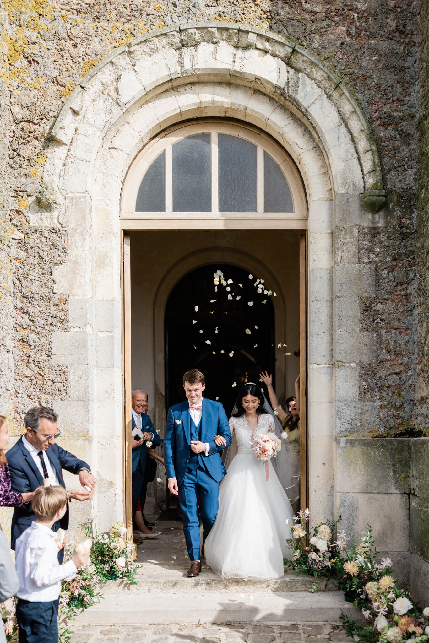 Couple exits stone chapel doorway in petal toss with blush and peach floral arrangements flanking arch