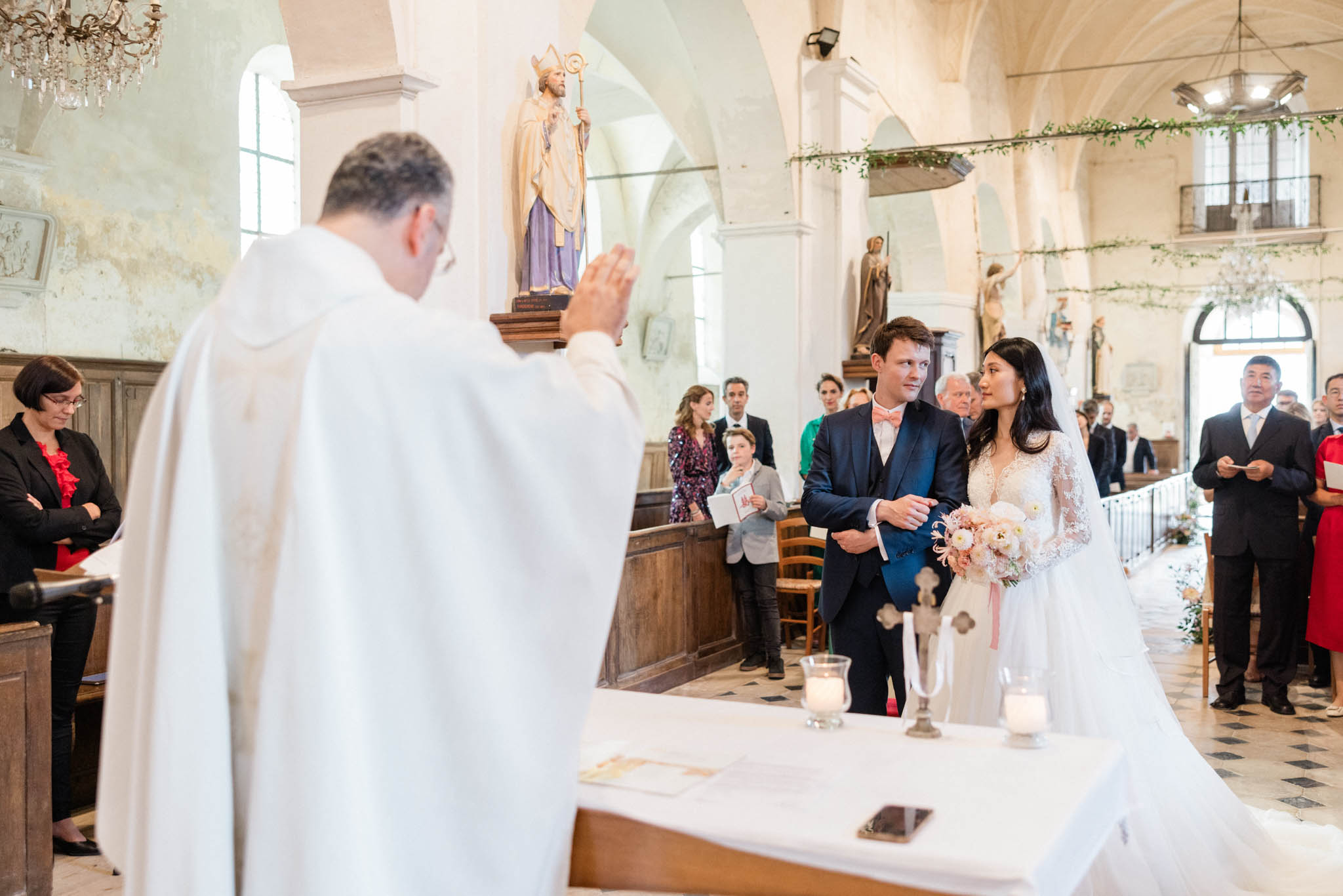 Catholic ceremony in vaulted chapel with priest blessing couple, greenery garlands, and checkered floor