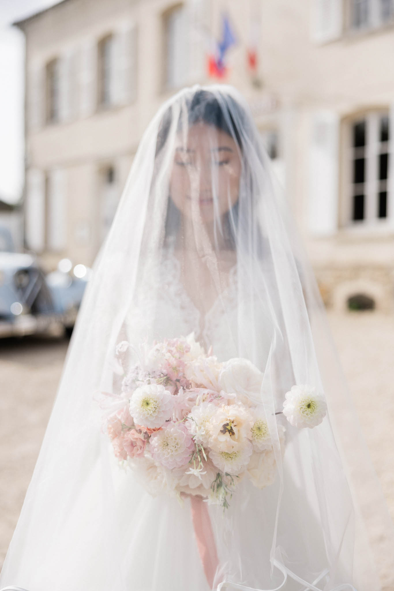 Bride in white ballgown and cathedral veil holding blush peony bouquet in limestone courtyard with vintage Citroen