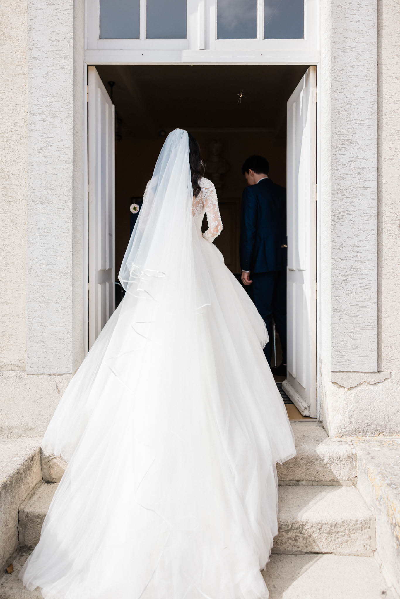 The bride and groom are captured from behind as they enter a building through large white double doors, likely at the start of or just after a ceremony. The bride wears a full ballgown-style white dress with a lace long-sleeve bodice and a voluminous multi-layered tulle skirt, paired with a long cathedral-length veil with a plain edge. The groom is dressed in a navy blue suit. The setting appears to be a formal stone venue, possibly a château or manor house, with classical stone pilasters flanking the doorway and stone steps leading to the entrance. The shot is a wide portrait taken from ground level behind the couple, emphasizing the dramatic train and veil of the bride's gown.