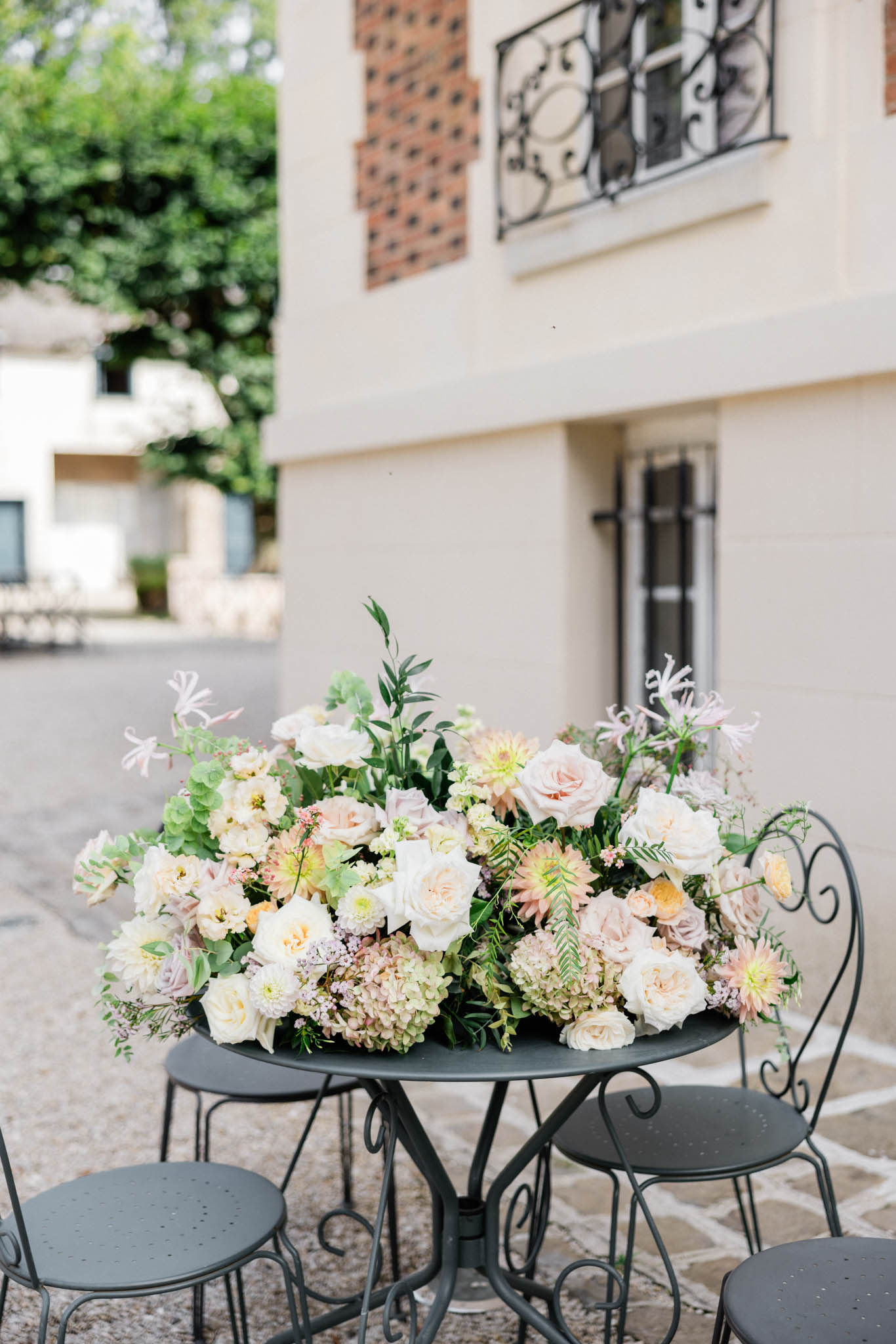 A close-up detail shot of a large floral centerpiece arrangement placed on a dark green wrought iron bistro table, set outdoors on a gravel and stone courtyard. The arrangement features blush roses, ivory garden roses, peach and yellow dahlias, soft green hydrangeas, pale pink nerine lilies, and mixed greenery including fern fronds and eucalyptus, creating a full, low, dome-shaped composition. The table is surrounded by matching dark green wrought iron bistro chairs, and the background shows a cream-rendered building facade with ornate black iron balcony railings and brick detailing. The overall floral palette is soft and muted — blush, cream, peach, soft green, and pale lavender — consistent with a classic French garden wedding aesthetic.