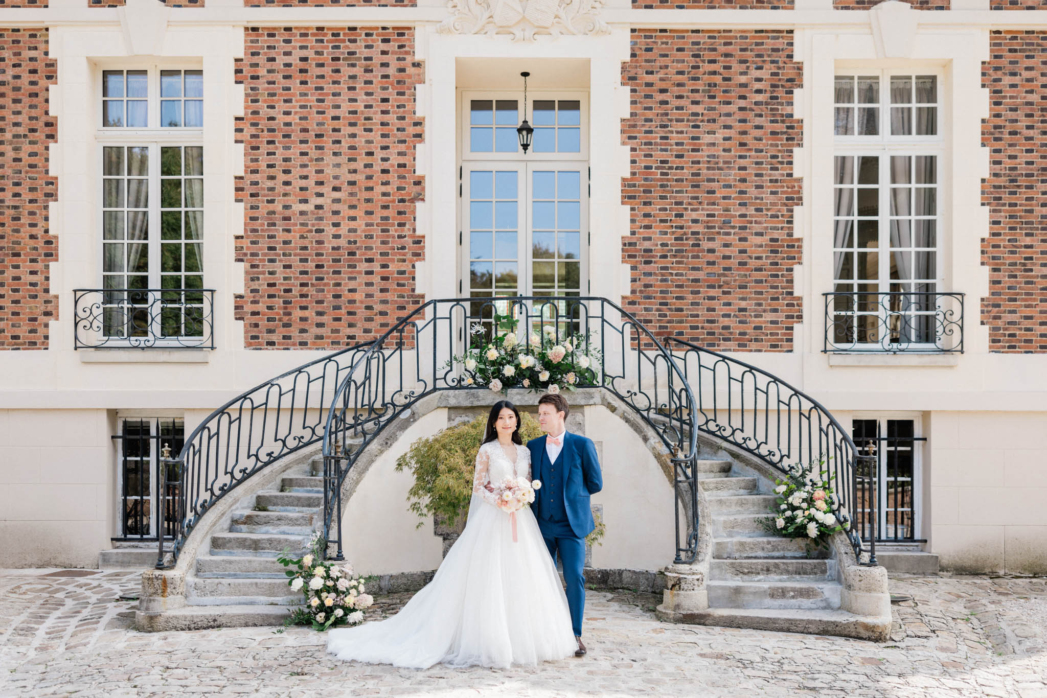 Couple before dual-curved stone staircase with blush and peach floral arrangements at red brick chateau