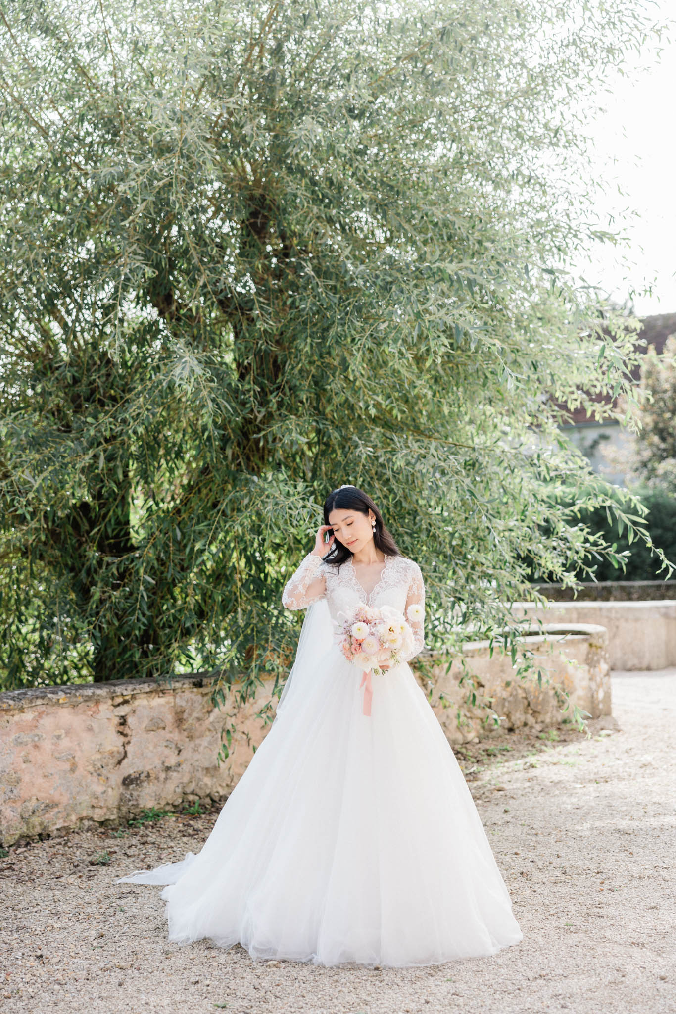 A bridal portrait taken outdoors on a gravel path, with the bride standing alone in front of a large weeping tree and a low stone wall. The bride wears a white ball gown with a lace V-neck bodice and long lace sleeves, with a full tulle skirt and a modest train. She holds a rounded bouquet of blush pink peonies, white ranunculus, and soft pink blooms tied with a blush satin ribbon, and she glances downward with one hand raised near her face. The overall styling is classic and romantic, with a soft, airy color palette of white and blush pink. Full-length portrait shot.