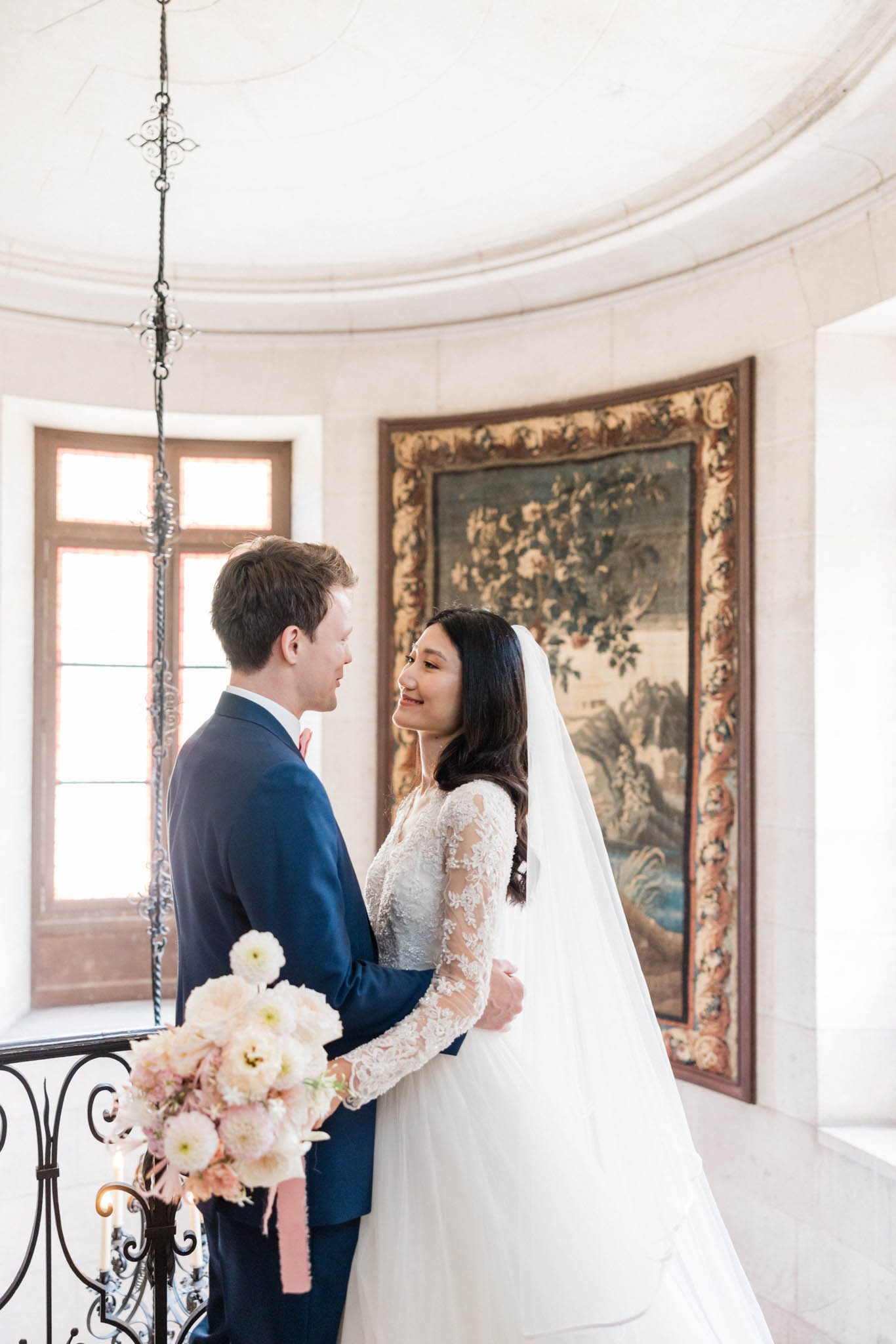 Bride and groom smiling on curved chateau staircase, bride holding cream and blush bouquet with pink ribbons