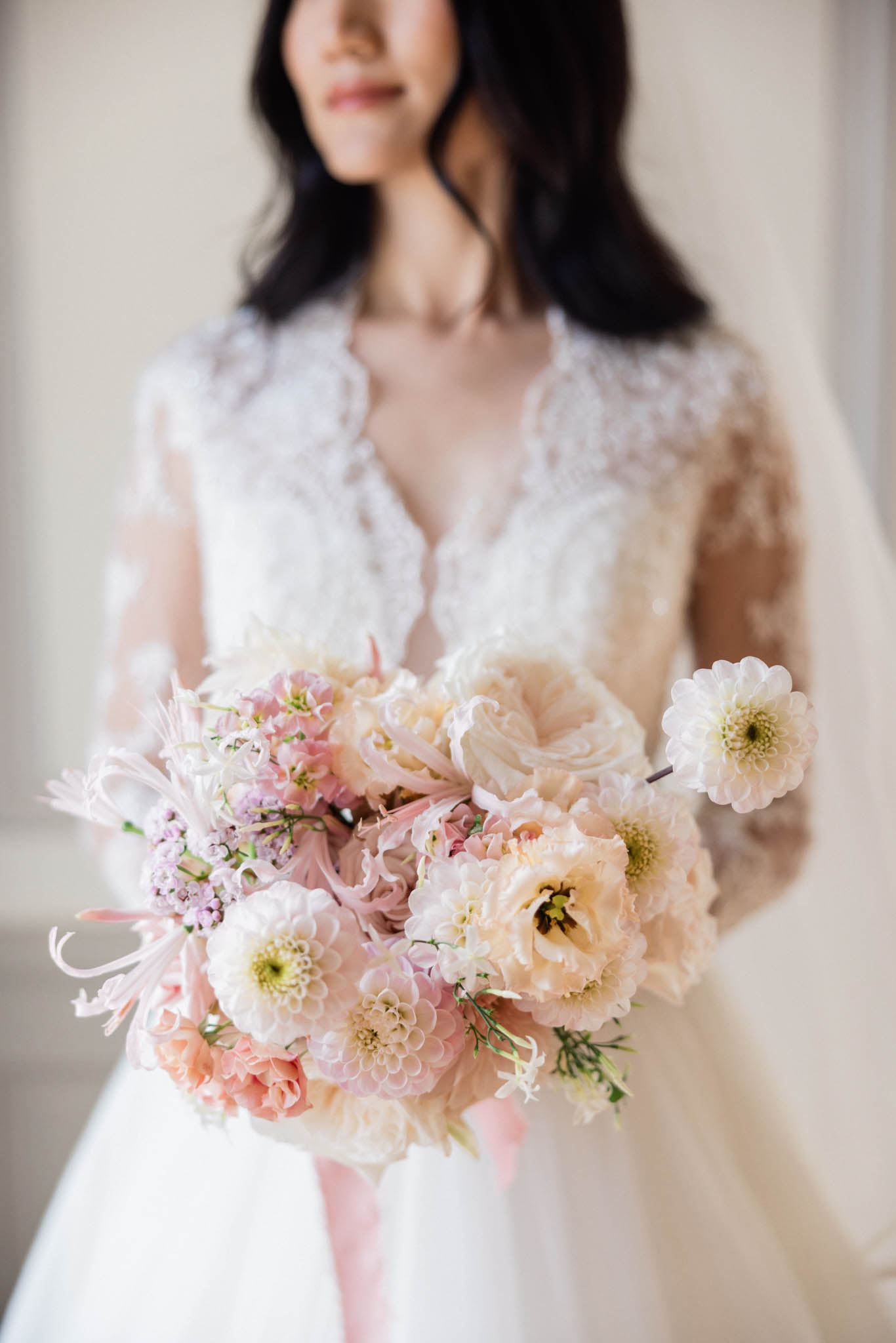 A close-up portrait of a bride holding her bouquet toward the camera in an indoor setting with a neutral, light-toned background. The bride wears a white long-sleeve lace gown with a deep V-neckline and floral lace appliqué detailing on the sleeves and bodice; her dark hair falls loosely around her shoulders. The bouquet is the primary focus of the image and features cream and blush dahlias, blush ranunculus, soft pink sweet peas, lilac wax flower clusters, and white spider-like blooms with wispy tendrils, composed in a rounded, garden-style arrangement. The composition uses a shallow depth of field, keeping the bouquet sharp while the bride's face and torso are intentionally soft-focused in the background.
