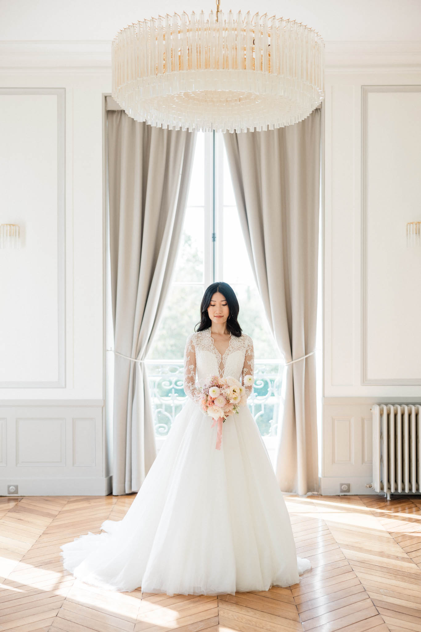 Bride in lace long-sleeve gown holding blush bouquet beneath crystal chandelier in panelled French interior