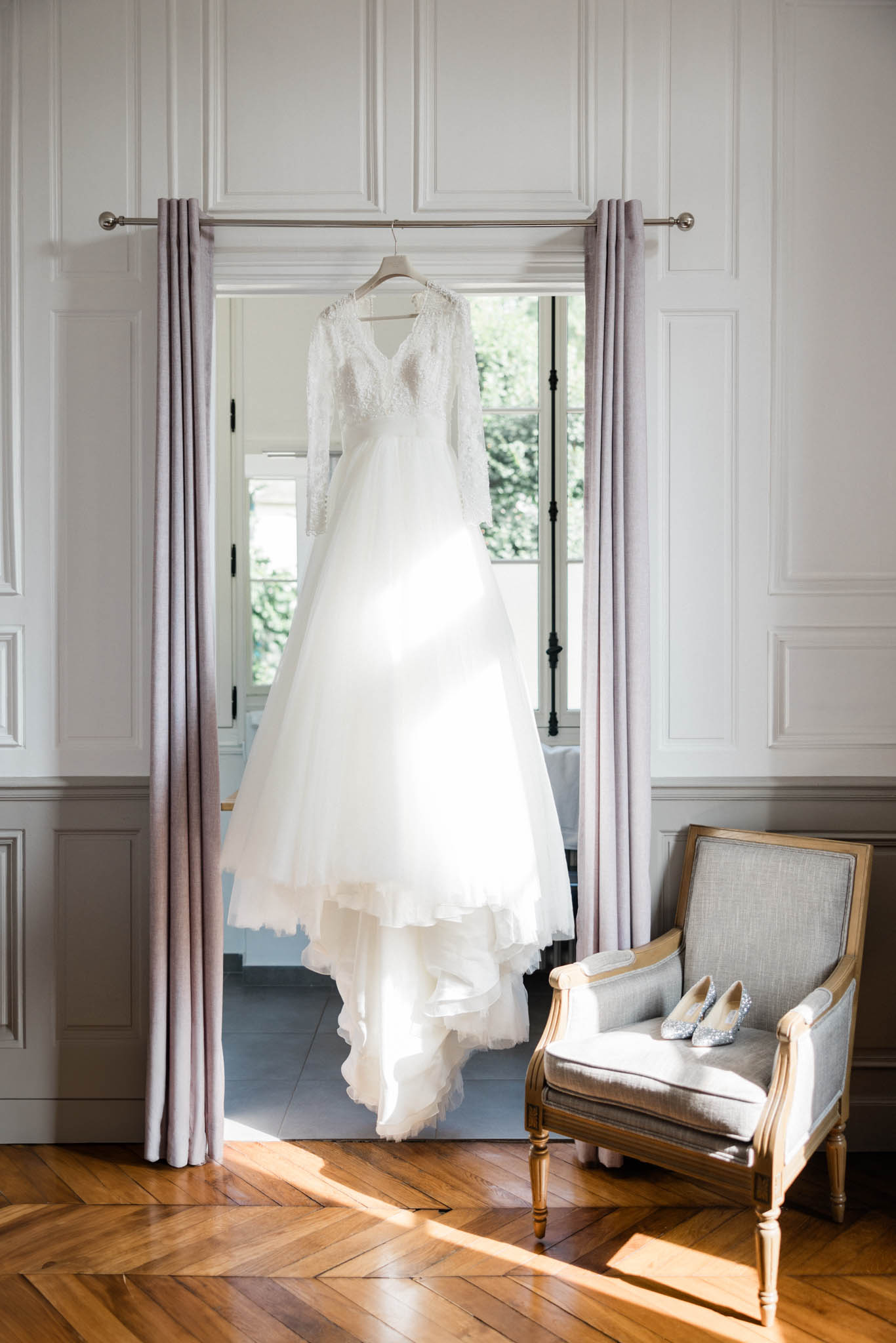Ivory lace-sleeve wedding gown hanging from curtain rod beside lavender drapes and silver bridal shoes on armchair