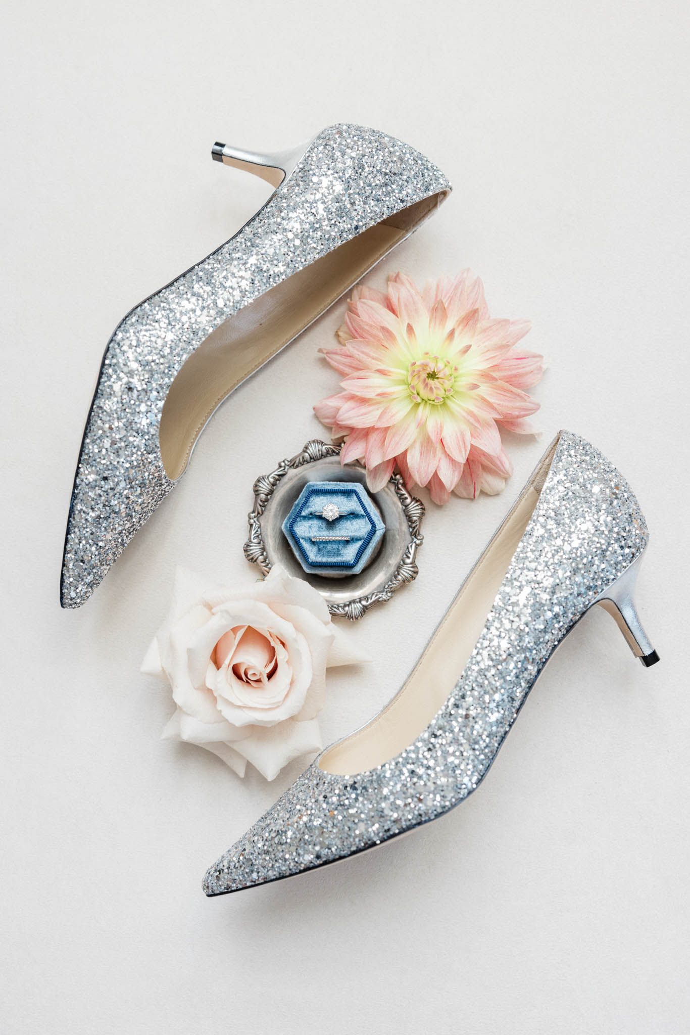 Flat lay of silver glitter heels, dusty blue velvet ring box, blush dahlia, and garden rose