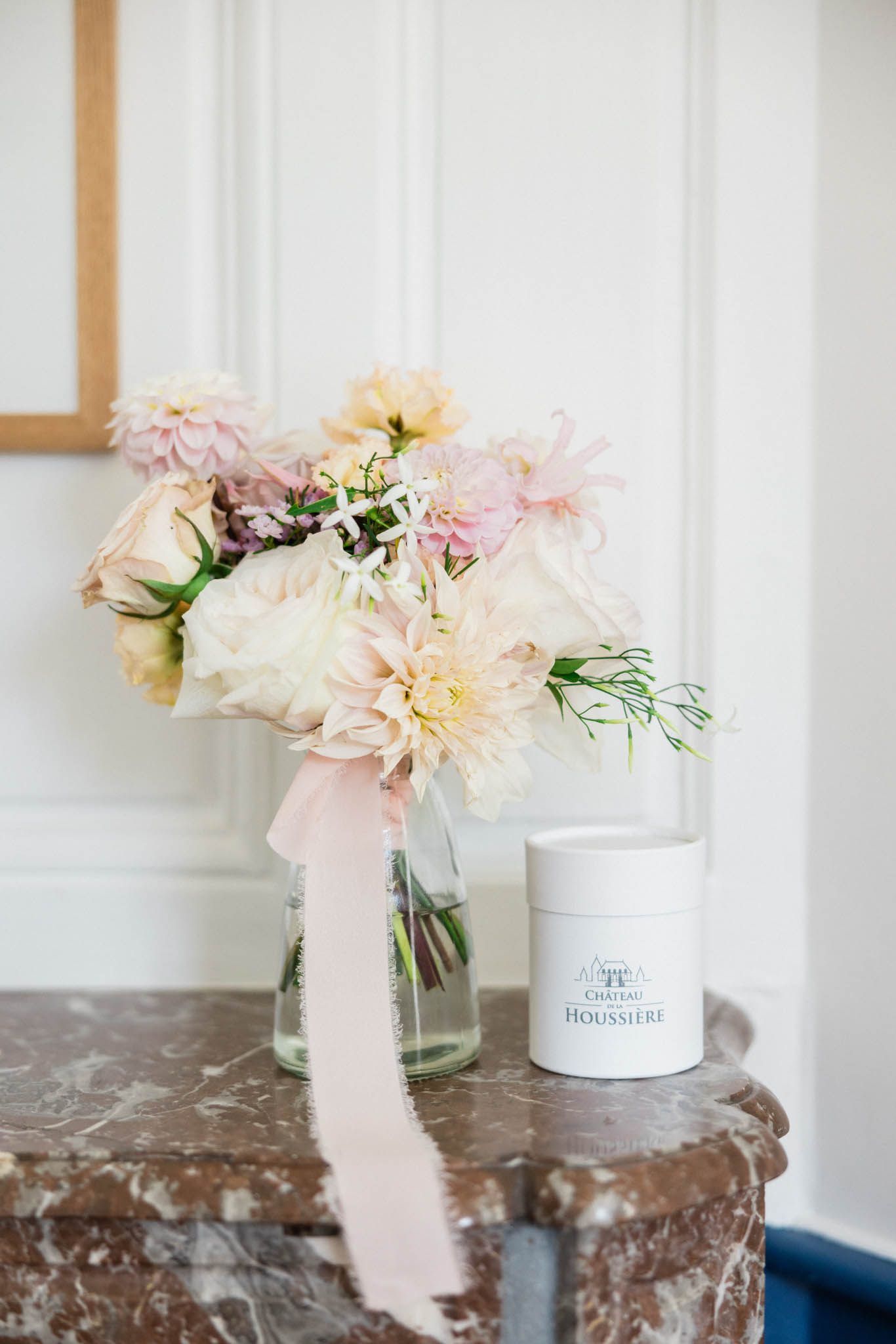 A close-up detail shot of a bridal bouquet placed in a clear glass vase on a marble mantelpiece inside a panelled white interior. The bouquet includes blush and cream dahlias, pale blush garden roses, small white star-shaped flowers, and delicate green filler stems, tied with a long frayed blush silk ribbon that drapes down the front of the vase. Beside the vase sits a white cylindrical branded candle or container printed with the Château de la Houssière logo. The overall decor palette is blush, cream, and soft pink, consistent with a classic romantic French château wedding aesthetic.