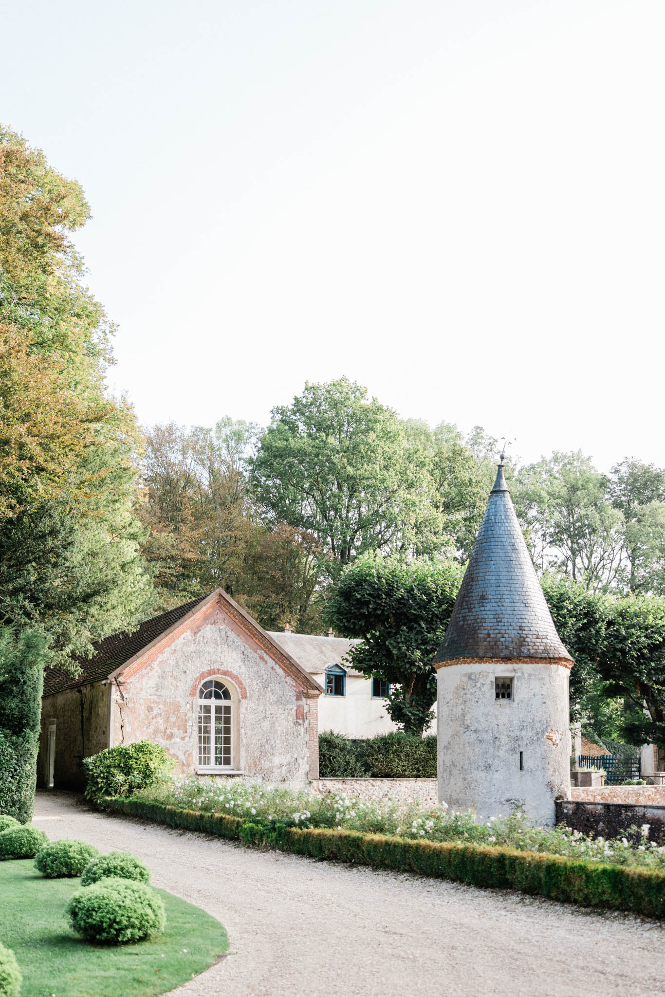 An outdoor wide shot of a French château estate's outbuildings and grounds, showing no wedding party or guests. The scene features a weathered stone and brick outbuilding with an arched white-framed window alongside a round tower with a dark slate conical roof, characteristic of French Renaissance architecture. The foreground includes a gravel driveway bordered by neatly clipped low boxwood hedges, rounded topiary shrubs on a lawn, and a bed of small white flowering plants lining the path. The image is shot in soft, bright natural light with an airy, slightly overexposed tone. Potential venue feature image.