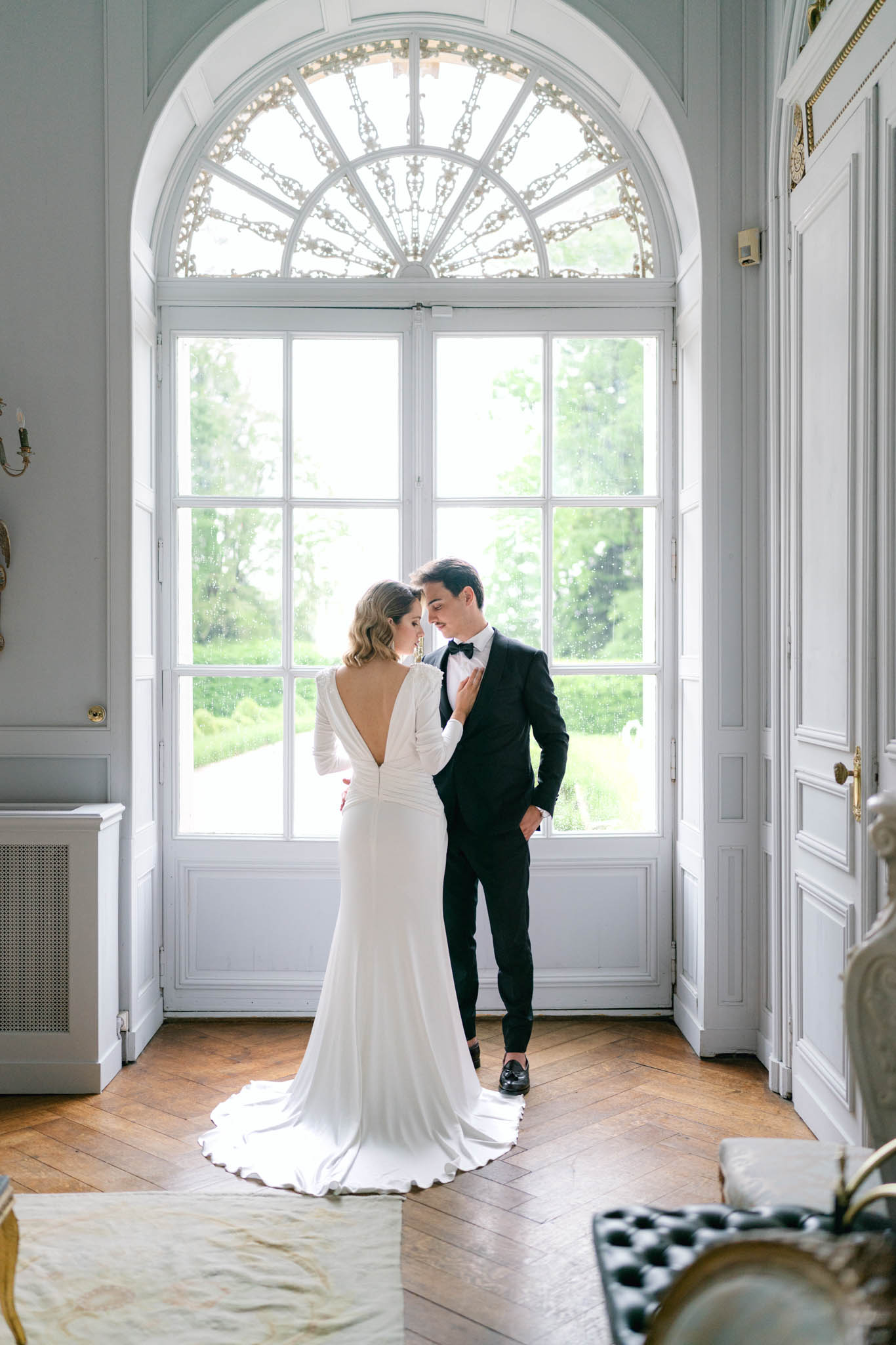 Bride and groom standing before arched fanlight door in chateau interior with white paneling and parquet floor