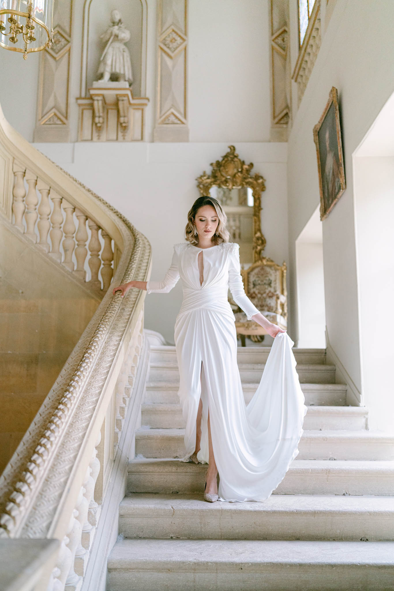 Bride in white long-sleeve draped gown descending a grand stone staircase inside a French chateau