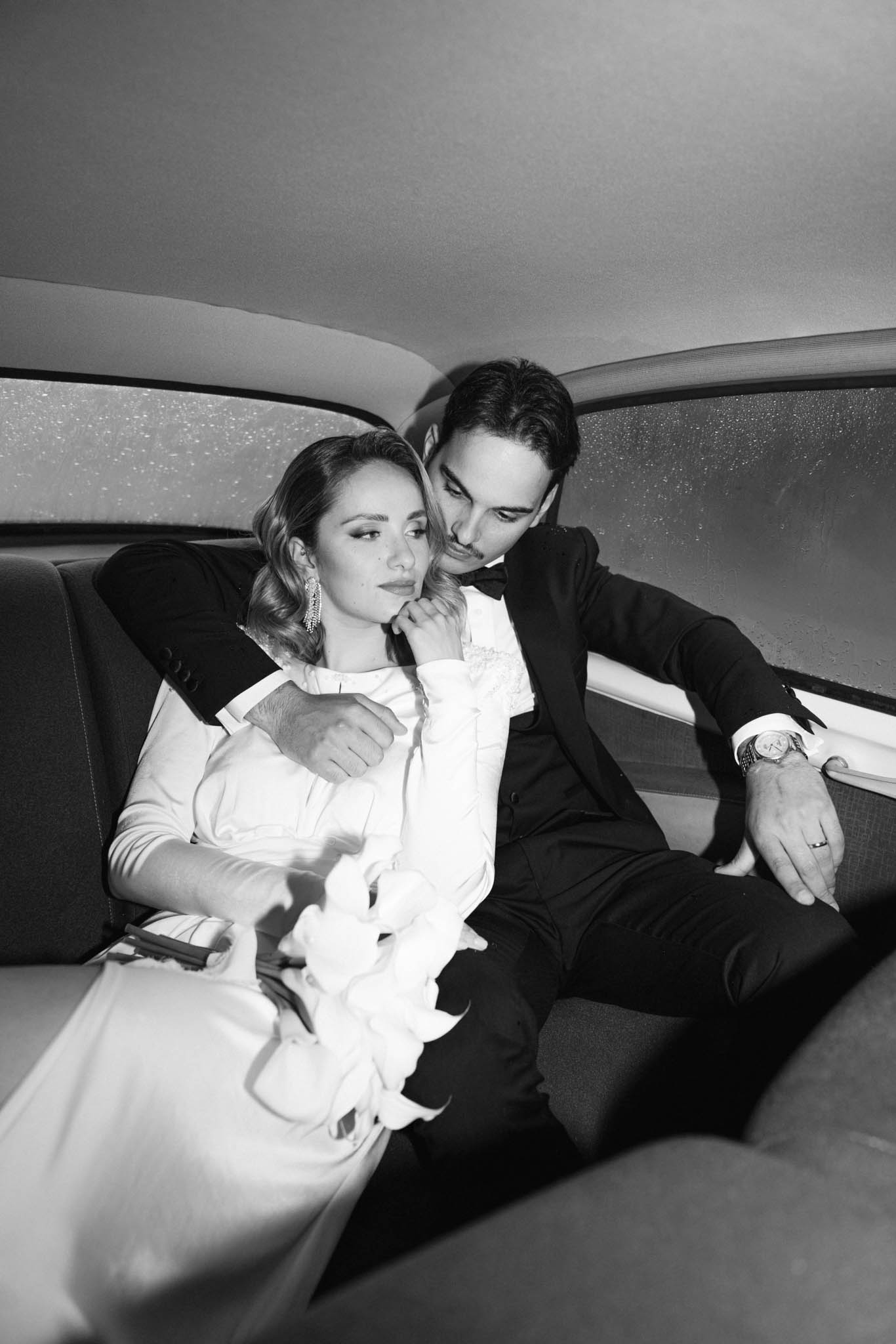 Black and white couple in vintage car backseat, bride holding white calla lily bouquet, groom in tuxedo