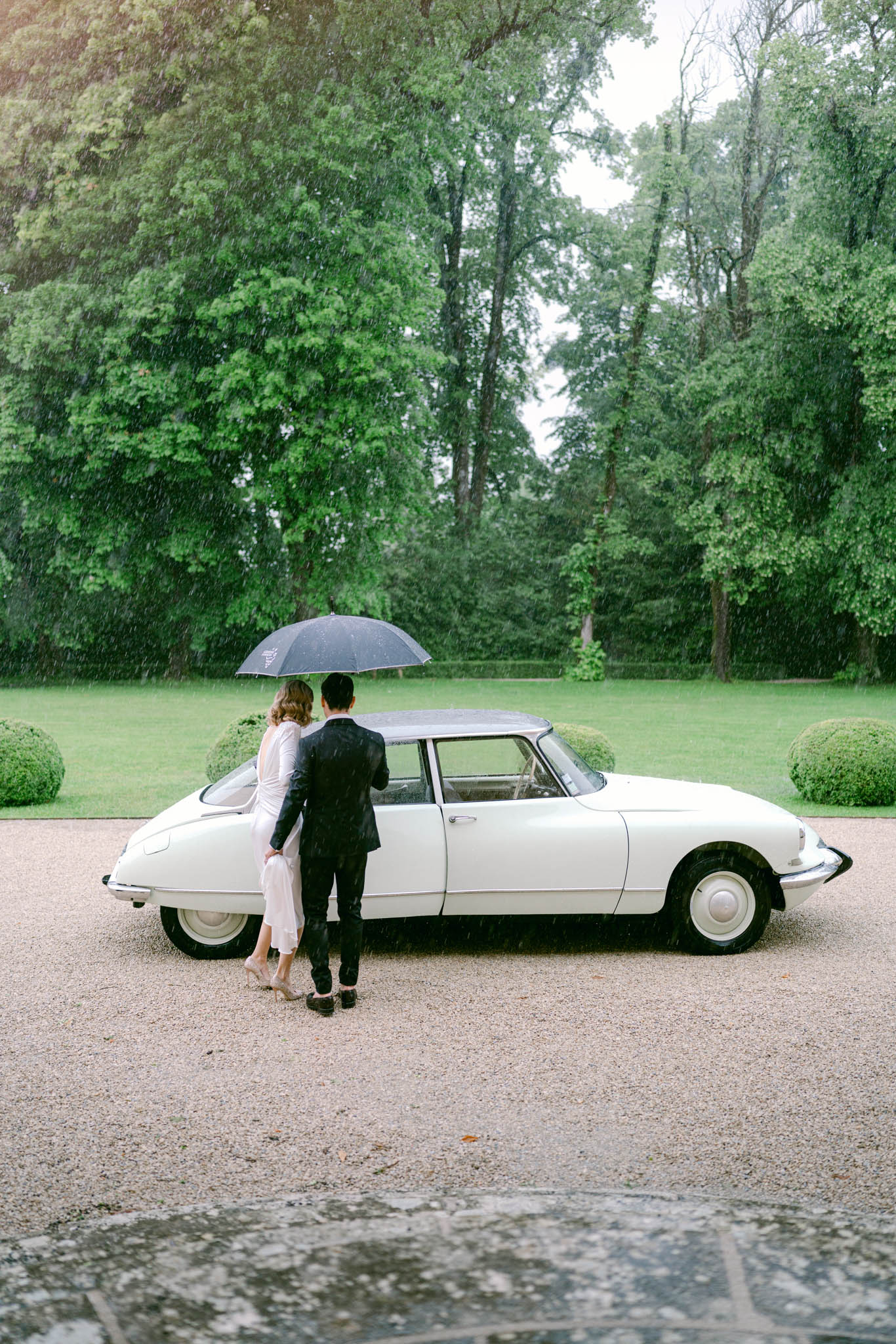 Bride and groom sharing umbrella beside vintage cream Citroen DS in rain on chateau gravel drive