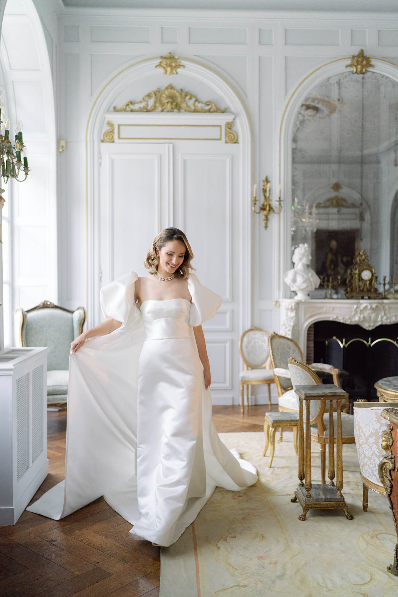 A bride stands alone in an ornate château salon, smiling down as she lifts the structured overskirt of her ivory satin wedding gown, which features dramatic oversized organza puff sleeves and a fitted column silhouette with a long train. She wears a pearl choker necklace and has her hair styled in a short, softly waved bob. The interior room is decorated in a classic French style with white-painted boiserie paneling trimmed in gold, arched doorways with gilded cartouche detailing, gold wall sconces, a carved white marble fireplace with a classical bust and ornamental clock on the mantel, and Louis XVI-style armchairs upholstered in pale sage green and gilt frames arranged on a cream area rug over herringbone parquet flooring. The portrait is a full-length medium shot taken in soft, diffused natural light. Potential venue feature image.