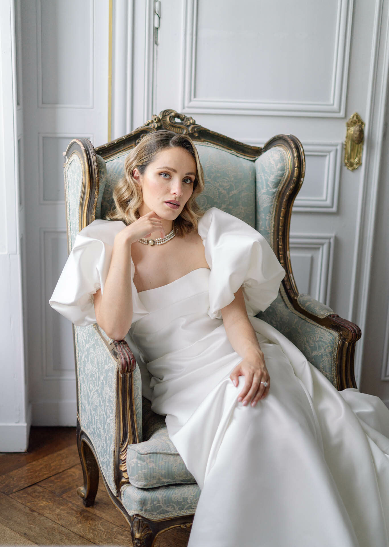 A bridal portrait taken indoors, showing the bride seated in a Louis XVI-style armchair upholstered in pale blue-green damask fabric with gold-painted carved wood framing. The bride wears a white satin gown with a square neckline and voluminous puff sleeves, accessorized with a multi-strand pearl choker necklace and a solitaire ring. Her blonde hair is styled in loose waves at shoulder length. The setting features white wood-paneled walls with classic boiserie molding and a gold door handle visible to the right, consistent with a French château or hôtel particulier interior. The shot is a medium portrait with natural, soft lighting, and the overall styling is classic French with a modern dress silhouette.