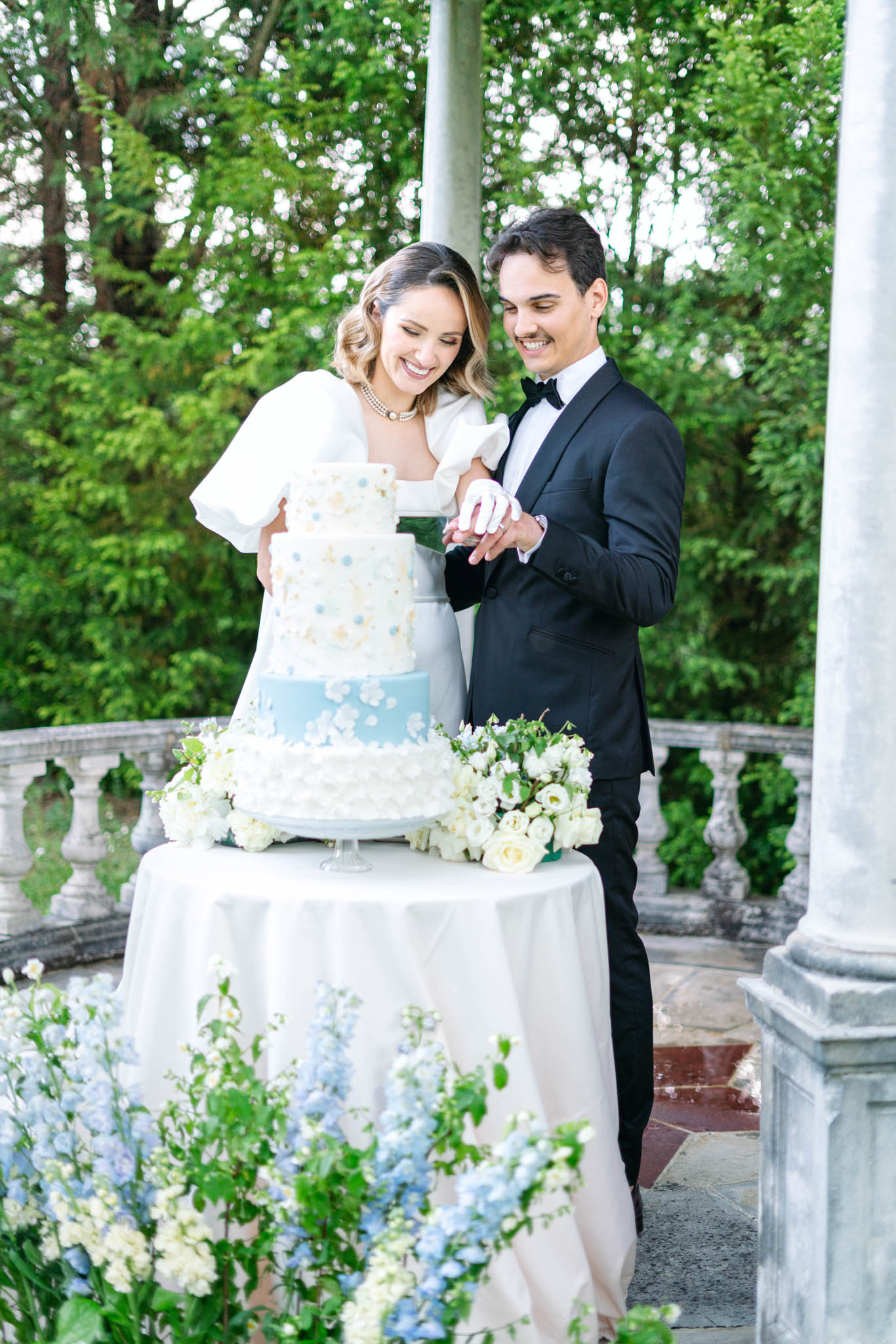 Couple cutting pale blue and ivory four-tier cake with sugar flowers on stone terrace
