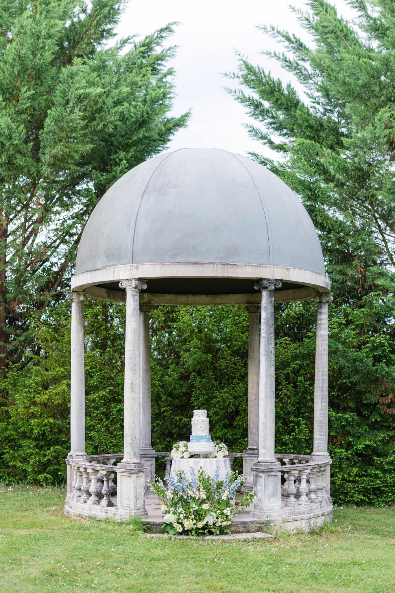 Blue and white tiered wedding cake in stone rotunda with hydrangea and delphinium arrangements