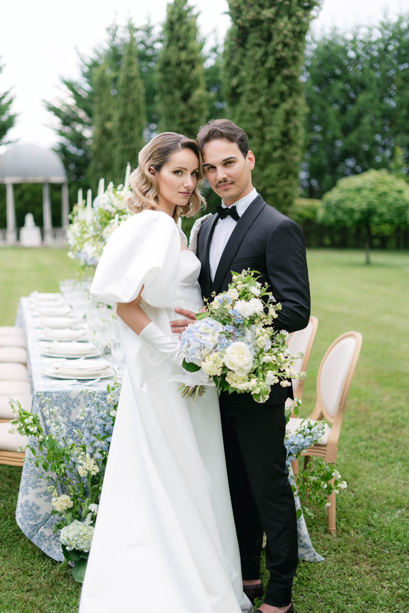 Bride in puff-sleeve gown and groom in black tuxedo beside a blue and white toile reception table with hydrangea arrangements