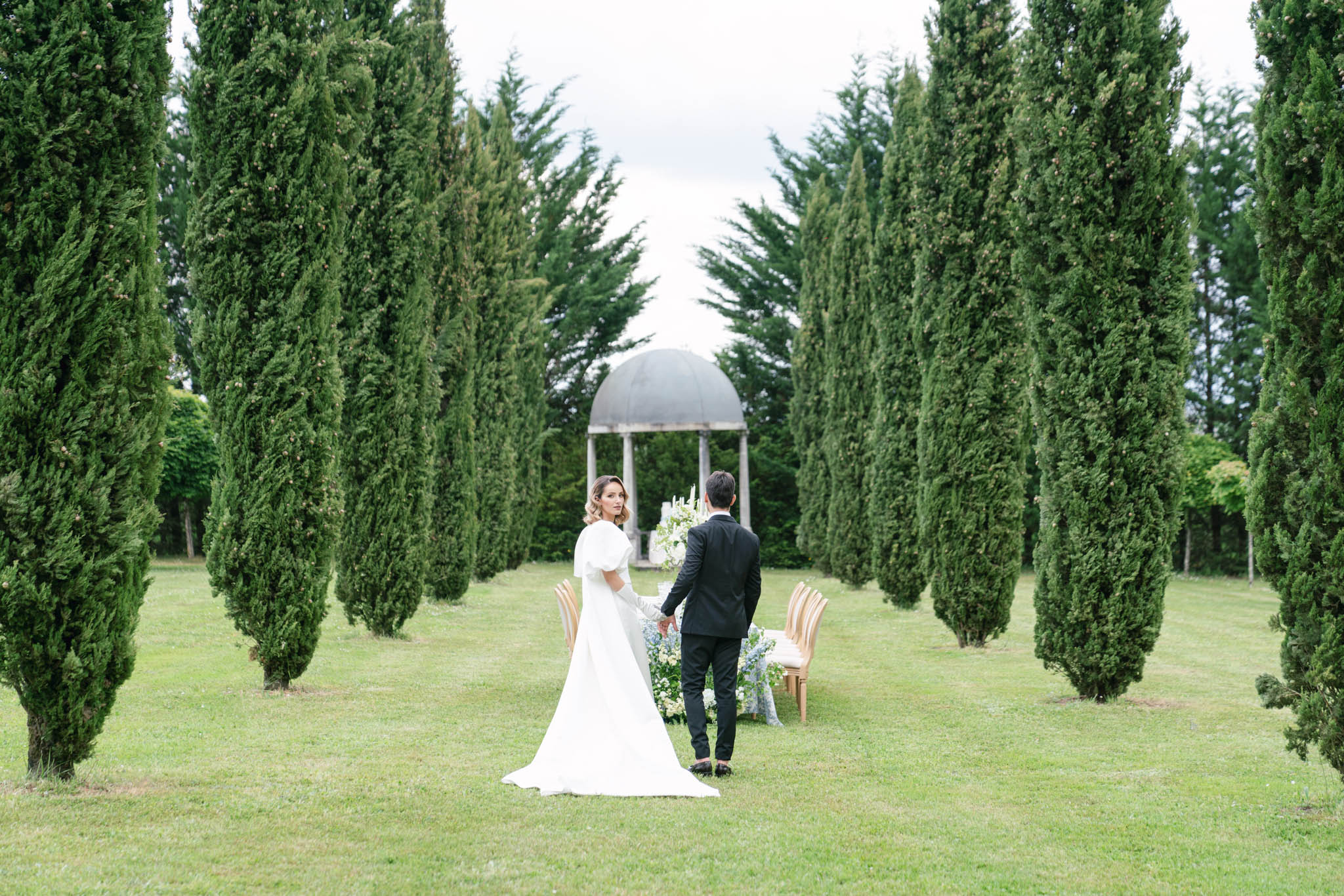 A couple walks hand-in-hand toward an outdoor ceremony setup in a formal garden allee lined with tall Italian cypress trees. The bride wears a white gown with puff sleeves and a long train, glancing back over her shoulder, while the groom is dressed in a black suit. Ahead of them, a stone domed rotunda serves as the ceremony backdrop, flanked by a floral arrangement of white and pale blue flowers with tall white taper candles, and gold Chiavari chairs positioned on either side. The styling is classic and refined, with a restrained white and soft blue floral palette. Wide shot taken from behind the couple, emphasizing the symmetry of the cypress allee and the rotunda as a focal point.
