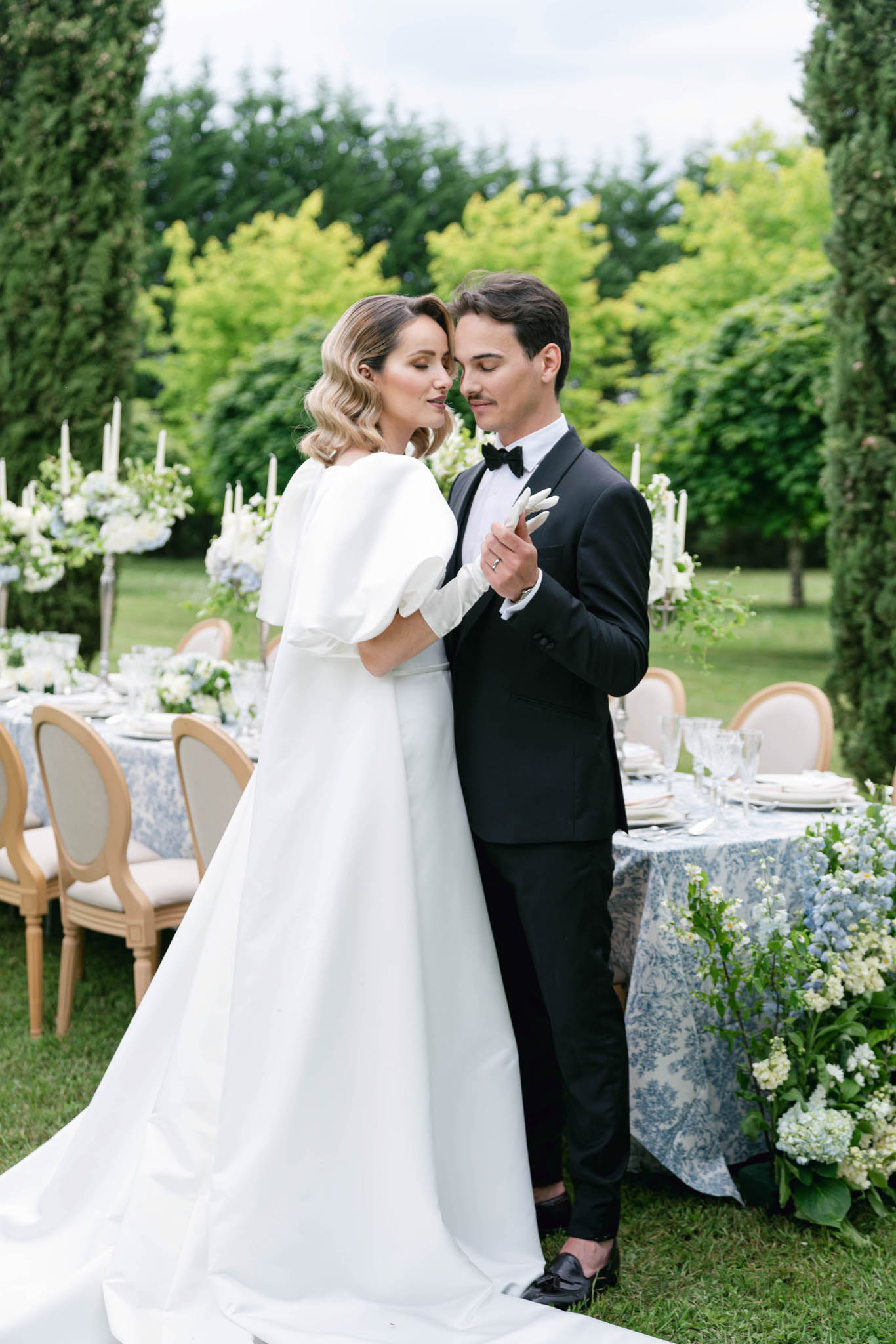 A couple portrait taken outdoors in a formal garden setting, with the bride and groom standing close together, foreheads nearly touching, in an intimate pose. The bride wears a white A-line gown with structured puff sleeves and a long train, paired with short white gloves and a soft wavy bob hairstyle; the groom wears a black tuxedo with a bow tie and black loafers. Behind them, a long outdoor reception table is dressed with a blue and white toile de Jouy tablecloth, white taper candles in tall candlesticks, stacked white plates, glassware, and low floral arrangements featuring pale blue delphinium, white blooms, and lush greenery; natural wood Louis XVI-style chairs line the table. The overall decor palette is blue, white, and natural wood, with a classic French-inspired styling theme, and the shot is a mid-length portrait framing both figures against the garden tableau.