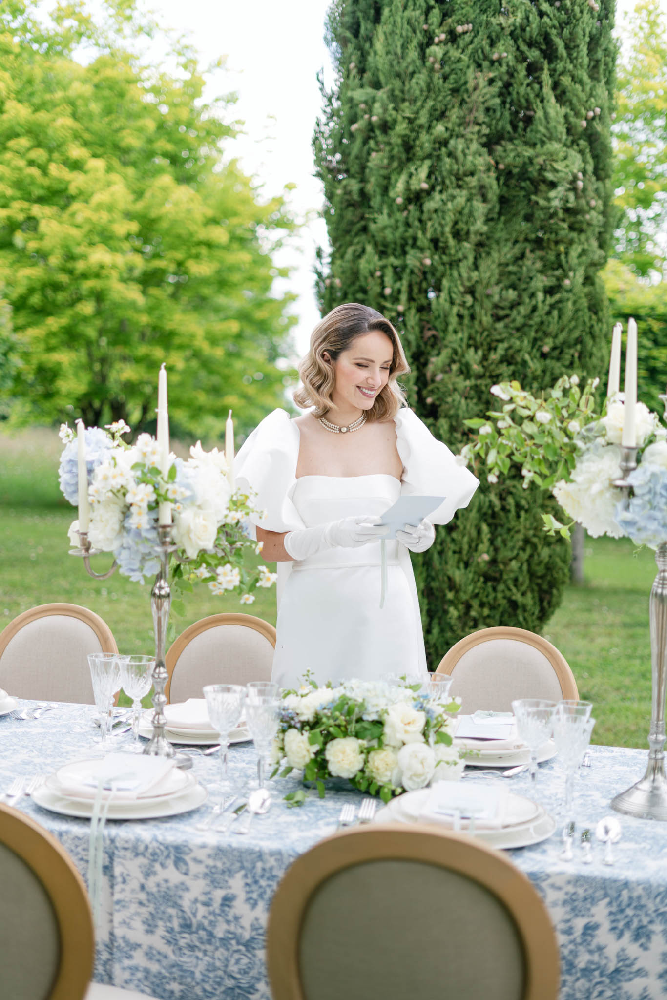 A bride stands at an outdoor reception table in a garden setting, smiling down at a small card she is holding. She wears a white strapless structured dress with detachable oversized puff sleeves, white gloves, and a pearl necklace. The table is dressed in a blue-and-white toile de Jouy printed linen and set with white china, crystal glassware, and silver cutlery. Centerpieces feature arrangements of ivory garden roses, white sweet peas, pale blue hydrangeas, and greenery displayed on tall silver candelabras with ivory taper candles, as well as a lower arrangement running along the center of the table. Chairs with natural wood frames and taupe upholstery surround the table. The overall décor palette is blue, white, and silver with a classic French formal styling. This is a medium portrait-style shot with the table in the foreground.