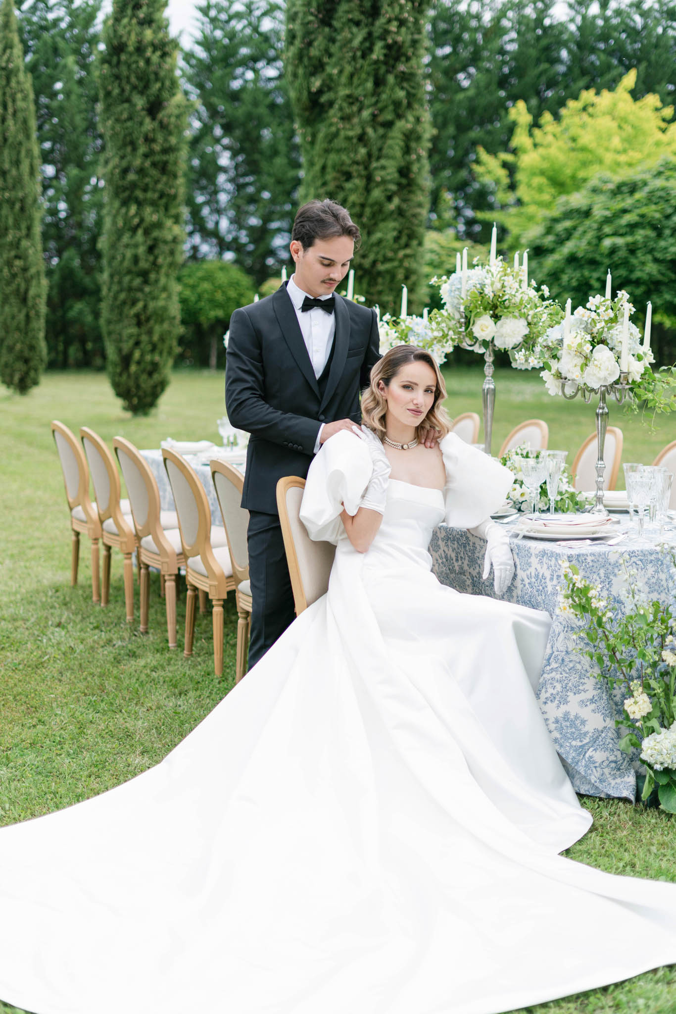 Bride in white ball gown and groom in tuxedo beside blue and white styled reception table with cypress trees