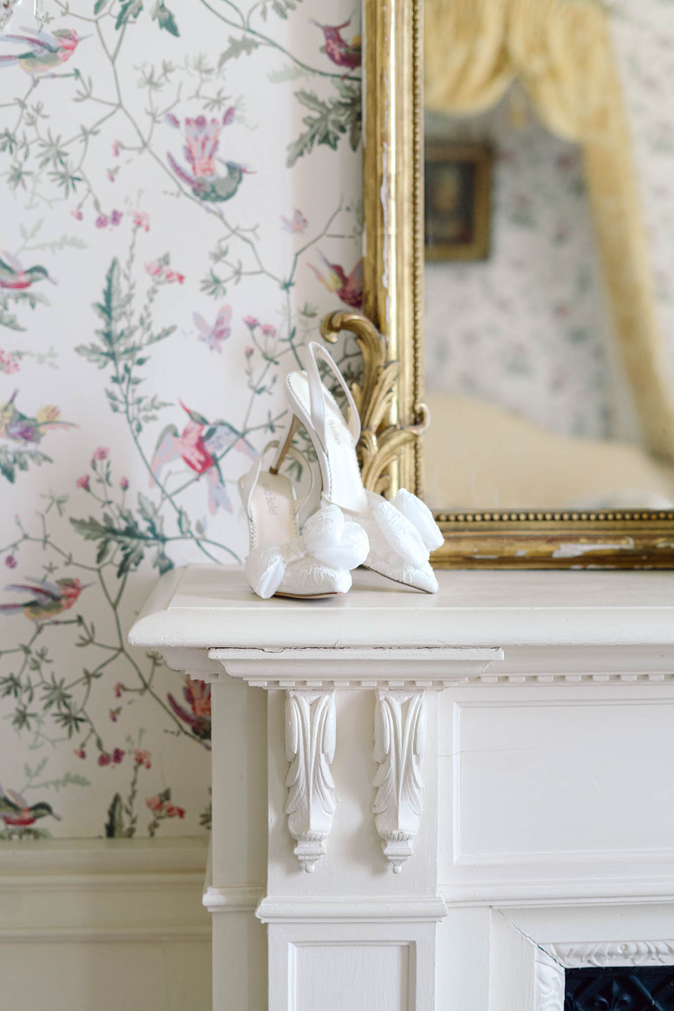 White slingback bridal heels with floral embellishments on an ornate fireplace mantel with gold rococo mirror