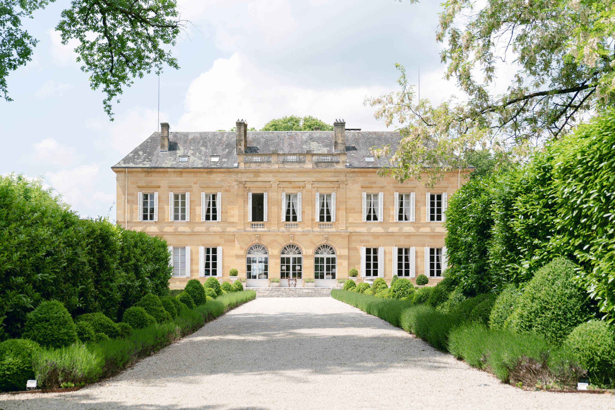 Honey limestone chateau with white shutters and mansard roof at end of lavender-lined gravel driveway