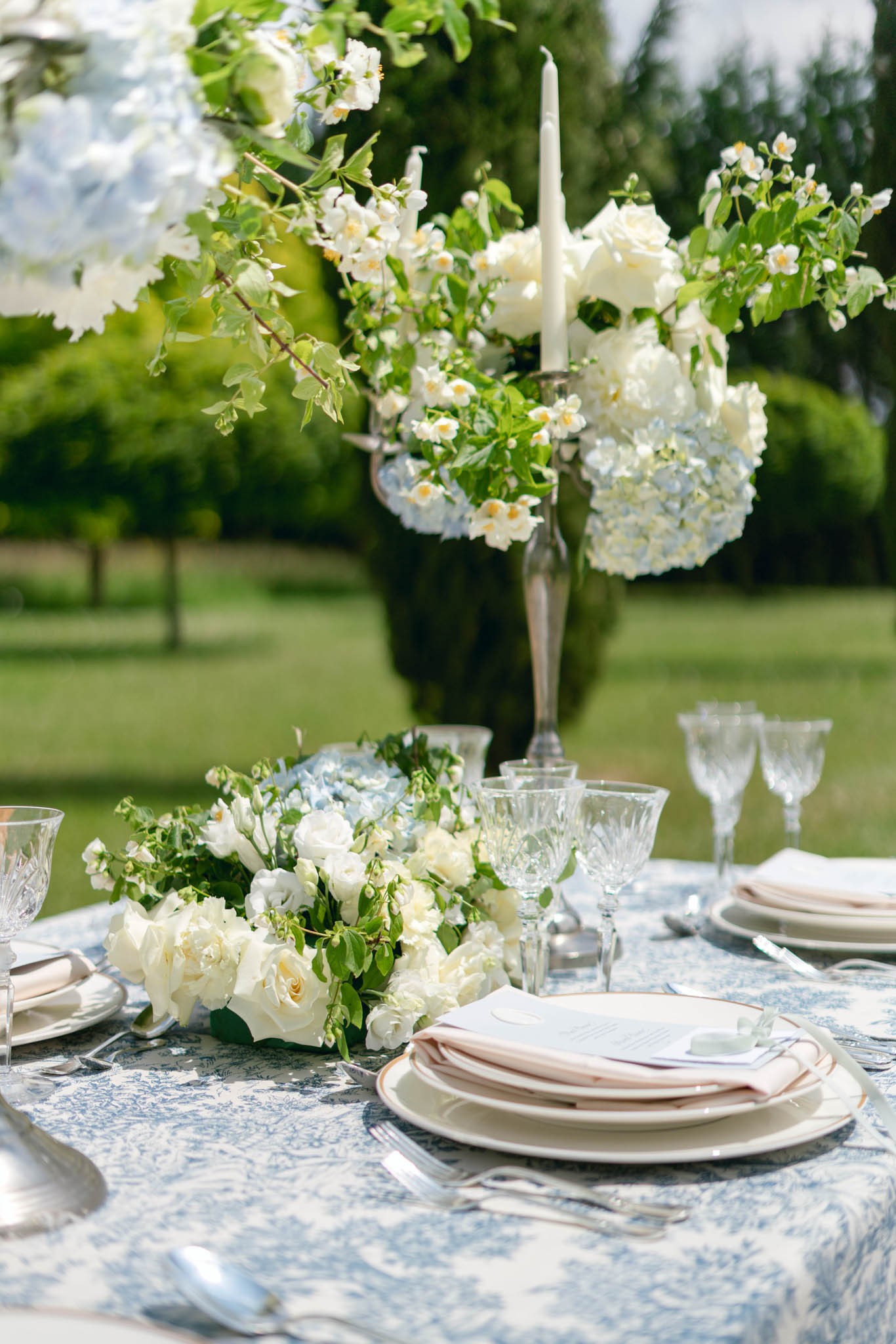 Garden reception table with blue toile de Jouy linen, gold-rimmed plates, cream roses, and silver candelabra with hydrangeas