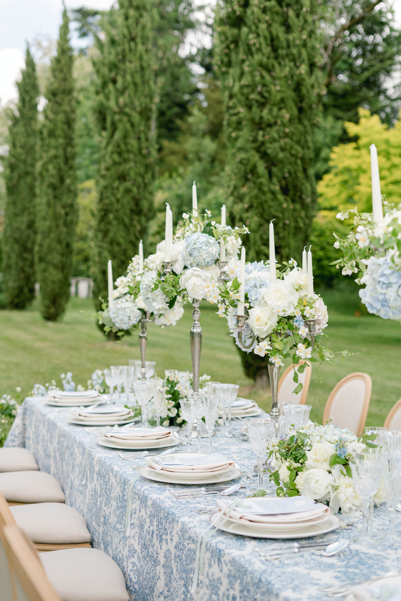 An outdoor wedding reception tablescape set in a formal garden with tall cypress trees in the background. The long rectangular table is dressed in a blue and white toile-patterned tablecloth and styled with stacked ivory plates, crystal glassware in varying heights, and silver flatware at each place setting. Tall silver candelabras serve as centerpieces, each adorned with white taper candles and arrangements of pale blue hydrangeas, white garden roses, white peonies, and trailing green foliage. A low floral runner of matching blooms, including white ranunculus and small blue accent flowers, runs along the center of the table between the candelabras. Natural wood and beige upholstered Louis XVI-style chairs line both sides of the table. The overall decor palette is blue, white, and silver with a classic French formal styling. Medium-distance angled shot.