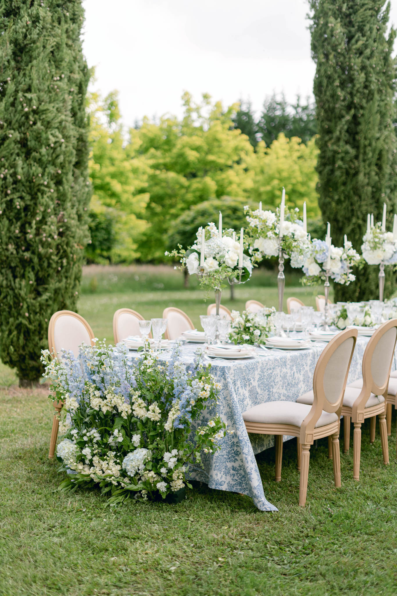 Long table in blue toile linen with blue delphinium cascade and silver candelabras between cypress trees