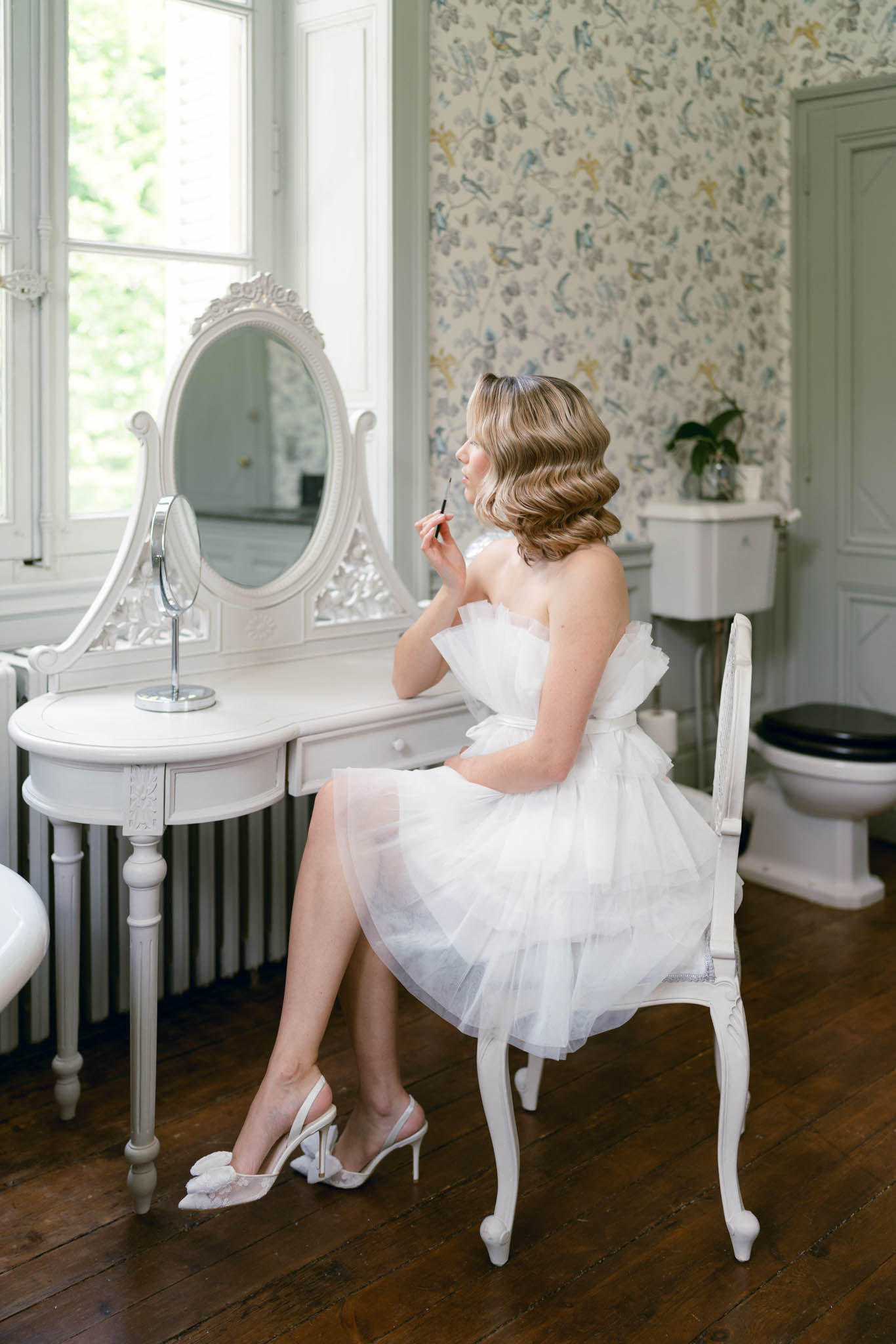 Bride in white tulle dress applying makeup at ornate vanity in chateau room with bird wallpaper