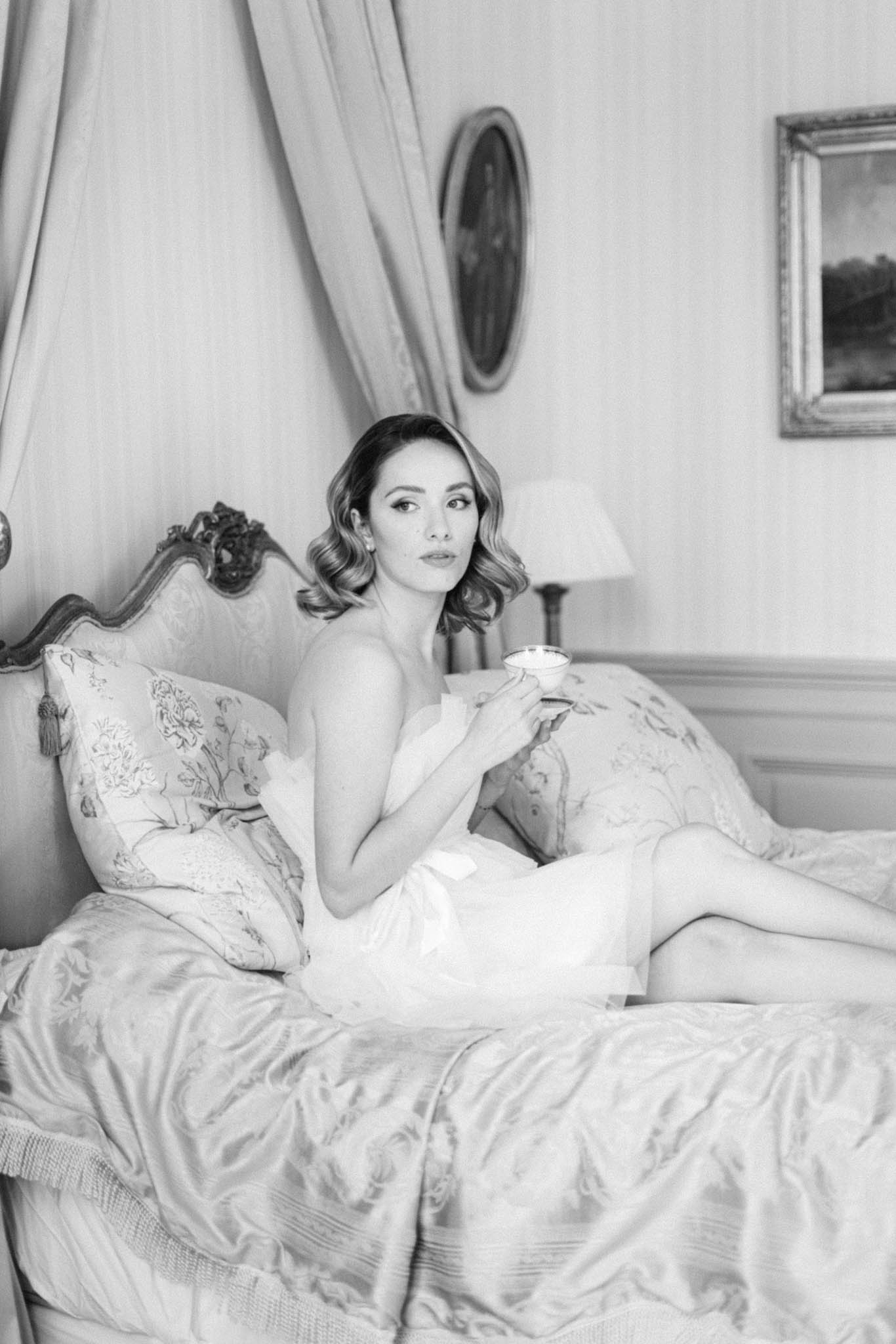 This black-and-white getting-ready portrait shows a bride reclining on an ornate carved wooden bed in what appears to be a château bedroom. She is wearing a strapless white silk slip dress or bridal robe and holds a fine china teacup, with her hair styled in soft vintage waves and her makeup fully done. The room features classic French interior details including jacquard floral bedding, floral-print cushions with tassel trim, draping fabric behind the headboard, gilt-framed paintings on the wall, and a bedside lamp. The image is a medium close-up portrait with soft, even tones and gentle contrast, conveying a classic, old-Hollywood morning-of bridal aesthetic.