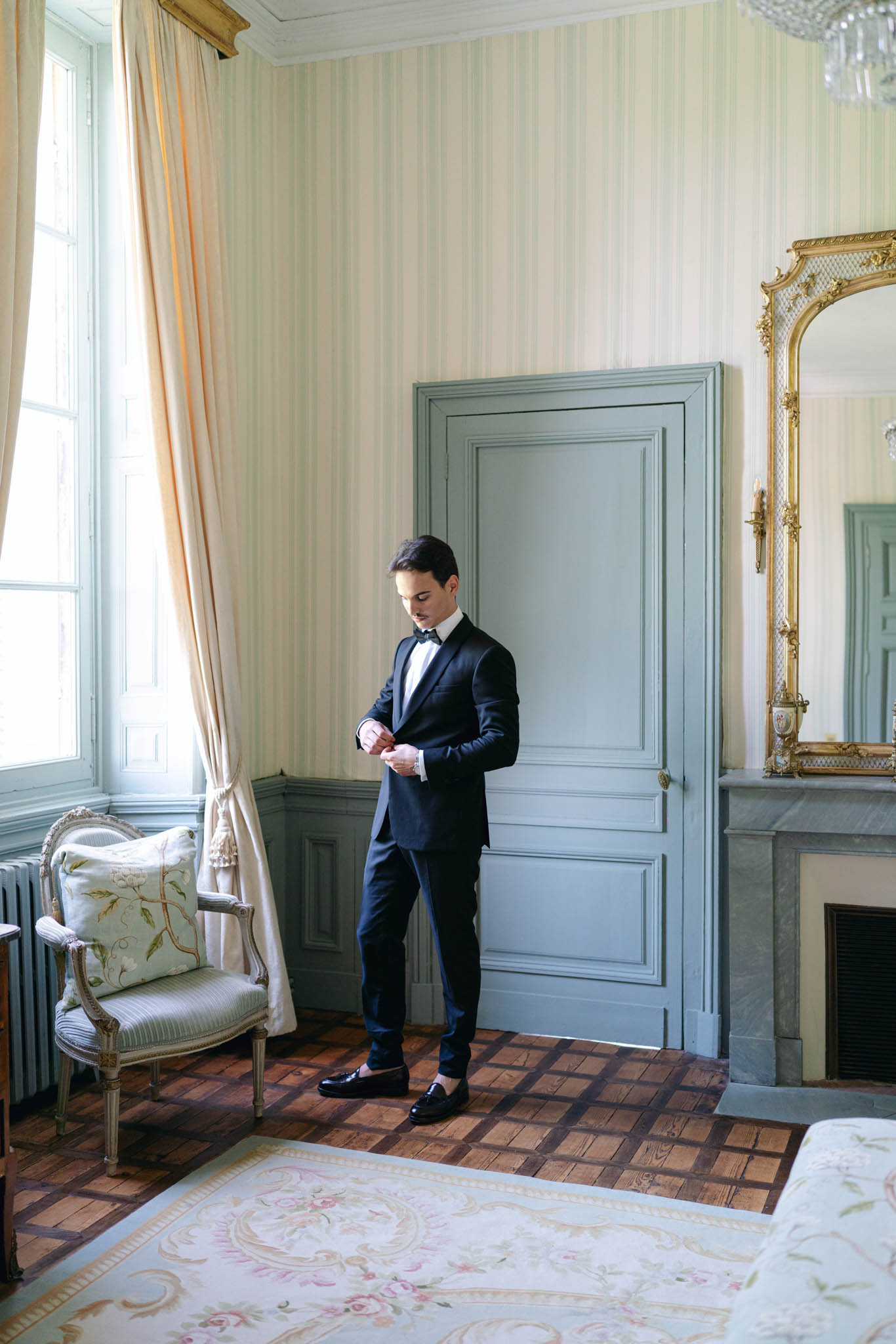 A groom getting ready in an ornate interior room, adjusting his jacket cuff or button while looking down. He is dressed in a navy tuxedo with a white dress shirt, black bow tie, and black patent leather loafers. The room features classic French château-style decor: sage-green painted panelled walls and doors, vertical stripe wallpaper, a parquet wood floor with a floral Aubusson-style area rug, a gilt-framed mirror above a marble fireplace, a Louis XVI-style armchair with a floral embroidered cushion, and a crystal chandelier partially visible in the upper right corner. Full-length curtains in pale peach frame tall windows. The composition is a full-length portrait shot with natural window light illuminating the scene.