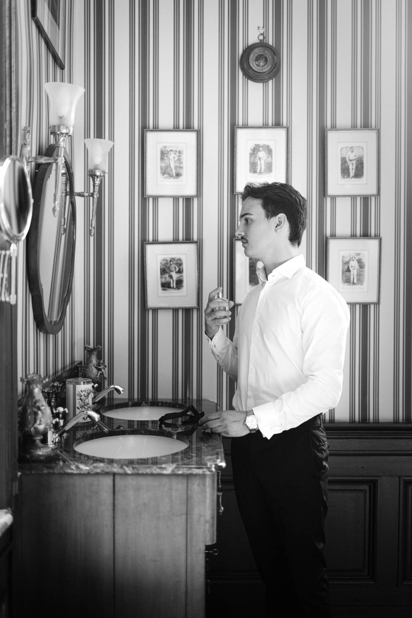Groom in white dress shirt applies cologne at marble vanity in ornate wallpapered bathroom in B&W
